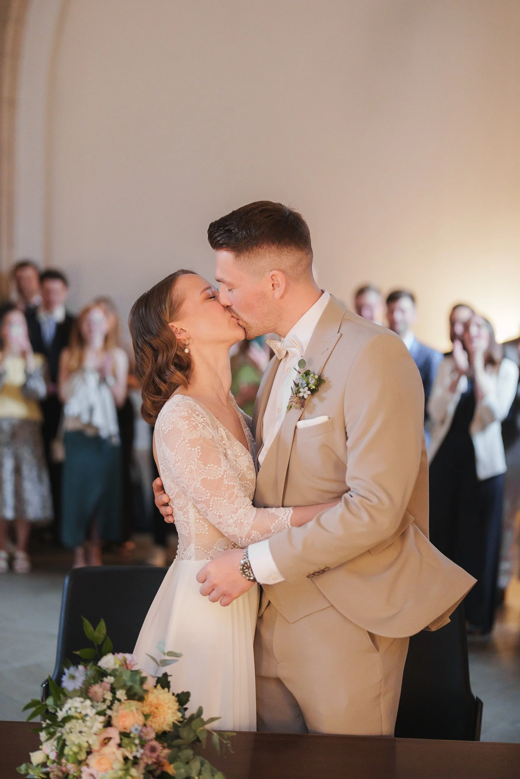 Ein Brautpaar küsst sich bei ihrer Hochzeit, im Hintergrund applaudierende Gäste.