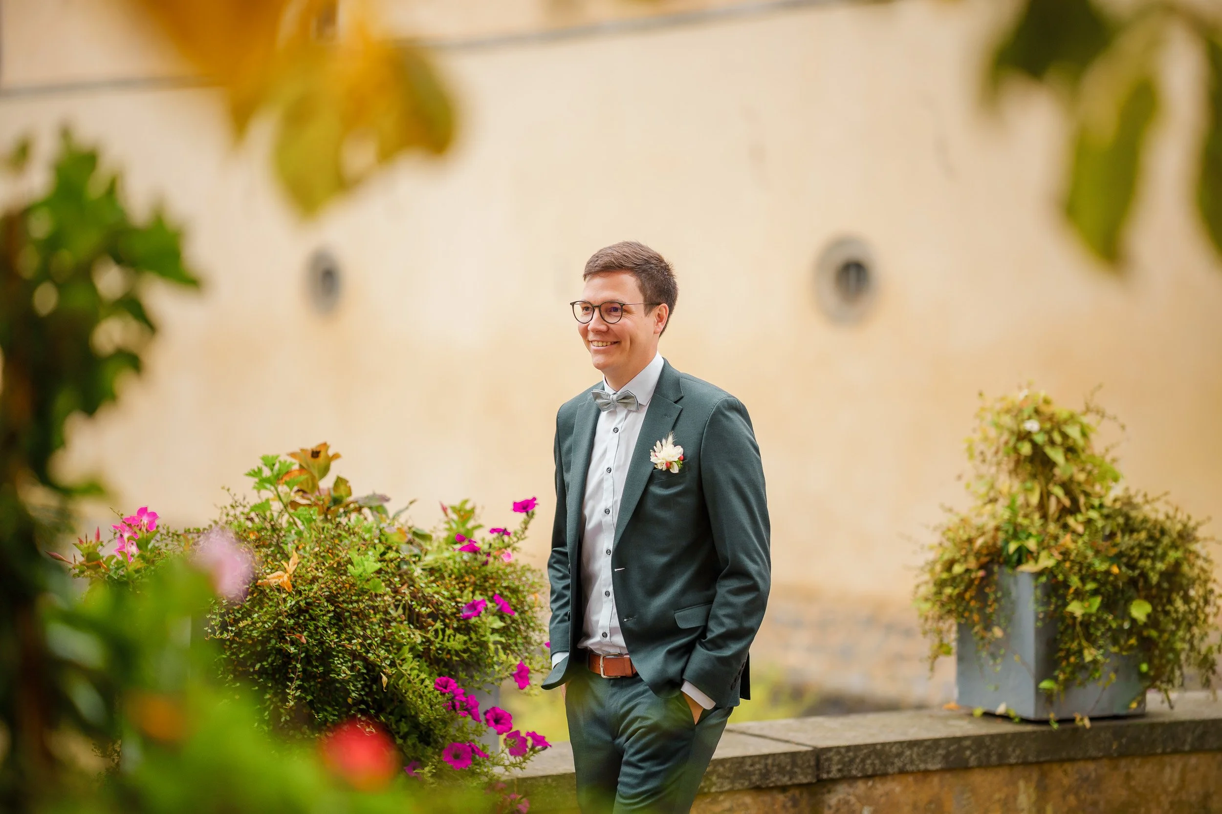Ein junger Mann in einem dunklen Anzug, weiße Hemd und Fliege lächelt, während er draußen neben blühenden Blumenkästen steht.