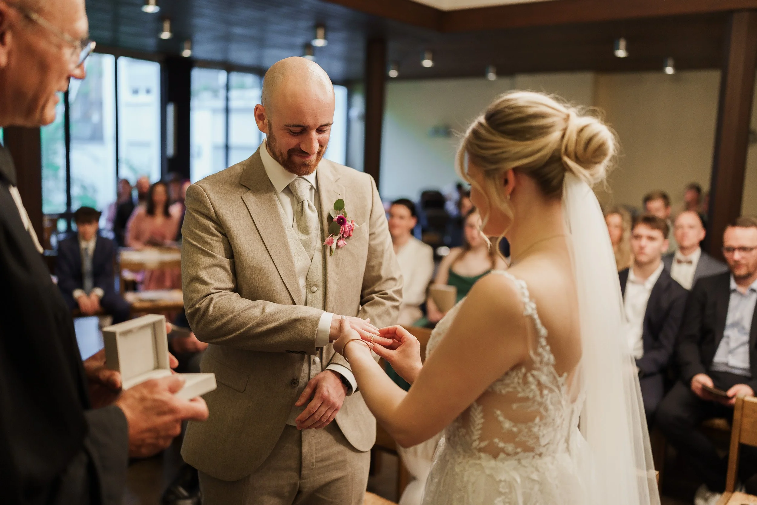 Ein Brautpaar bei der Trauung, der Bräutigam legt der Braut einen Ring an, im Hintergrund sitzen Gäste bei einer Hochzeitsfeier.