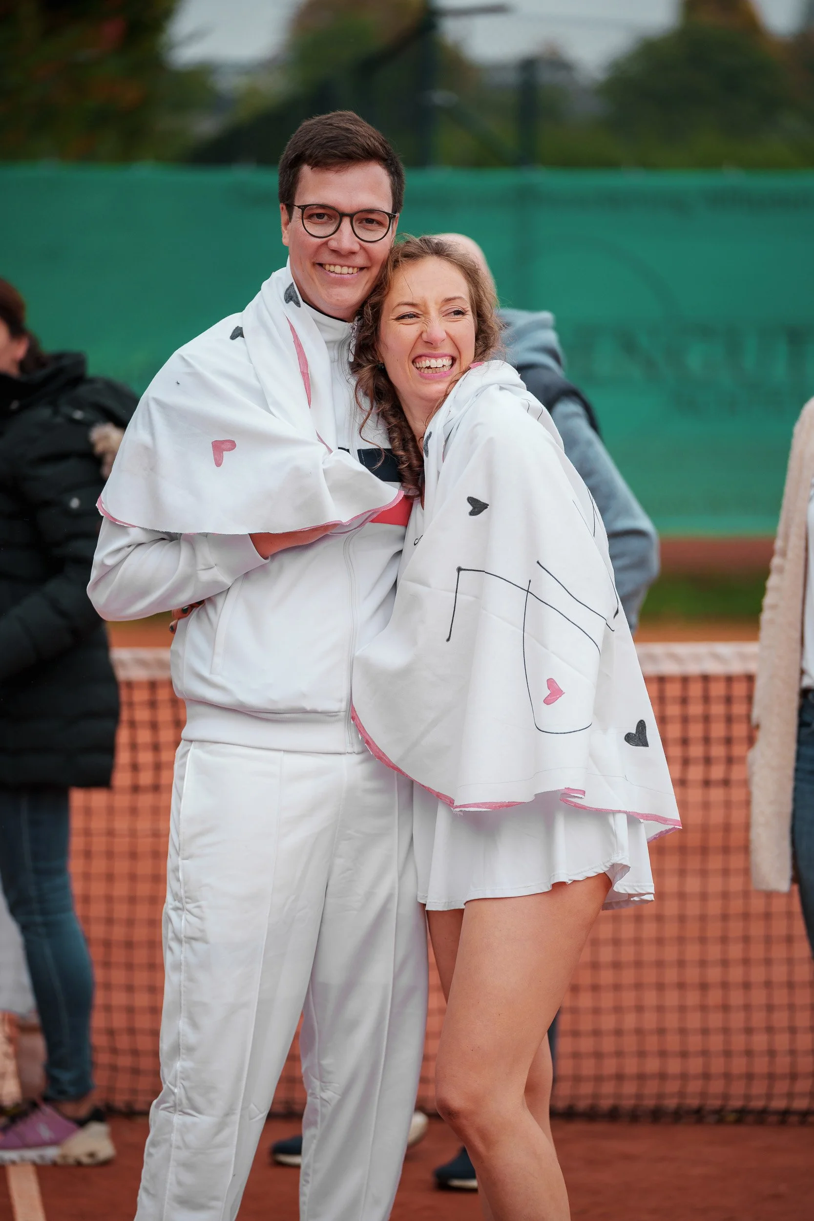 Ein Mann und eine Frau lachen und umarmen sich auf einem Tennisplatz, beide sind in weißen Sportoutfits, die Frau hat eine Decke mit Herzen um sich gewickelt.