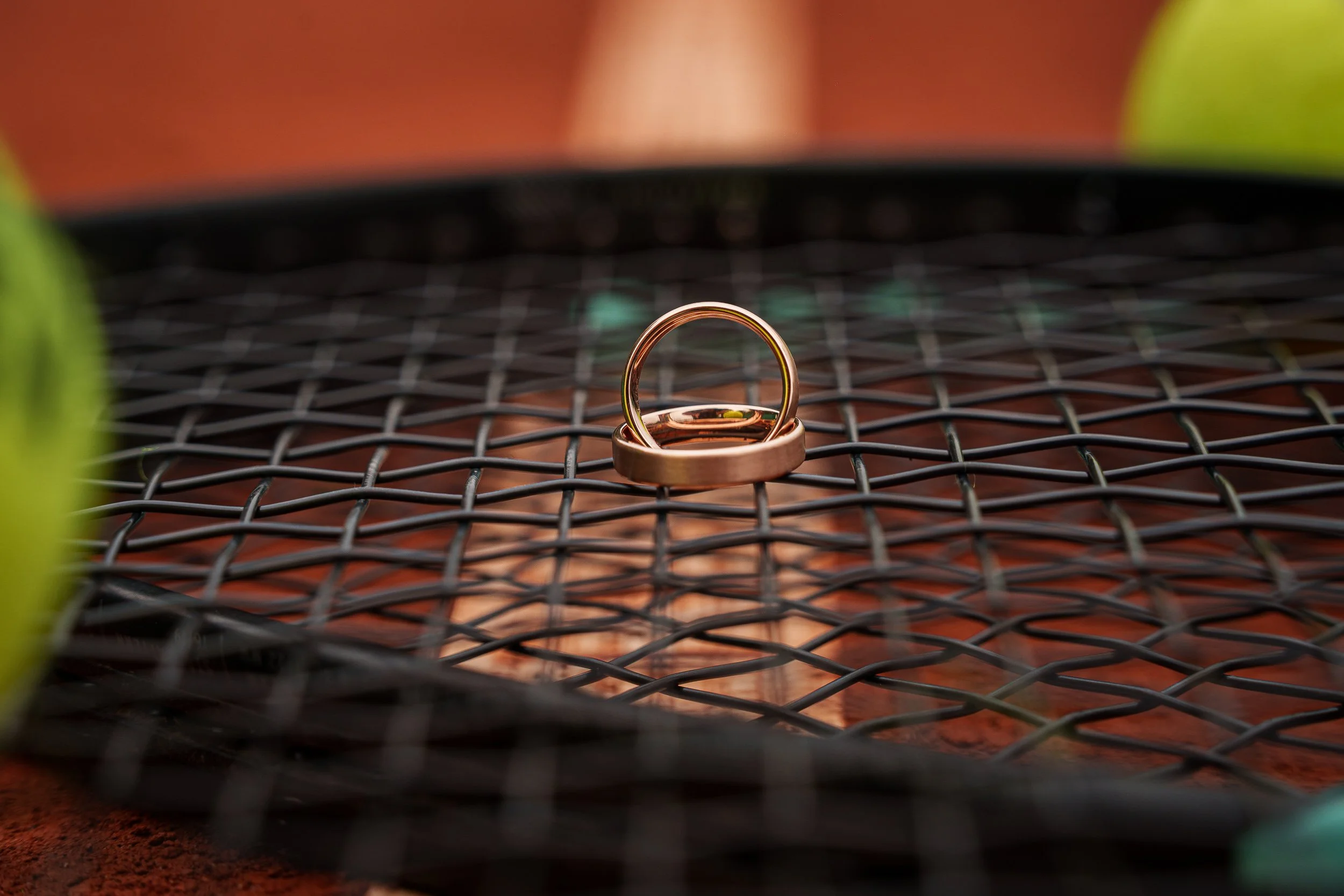 Zwei Goldringe liegen auf einer schwarzen Gittermatte, im Hintergrund ist eine Tennisracket sichtbar.