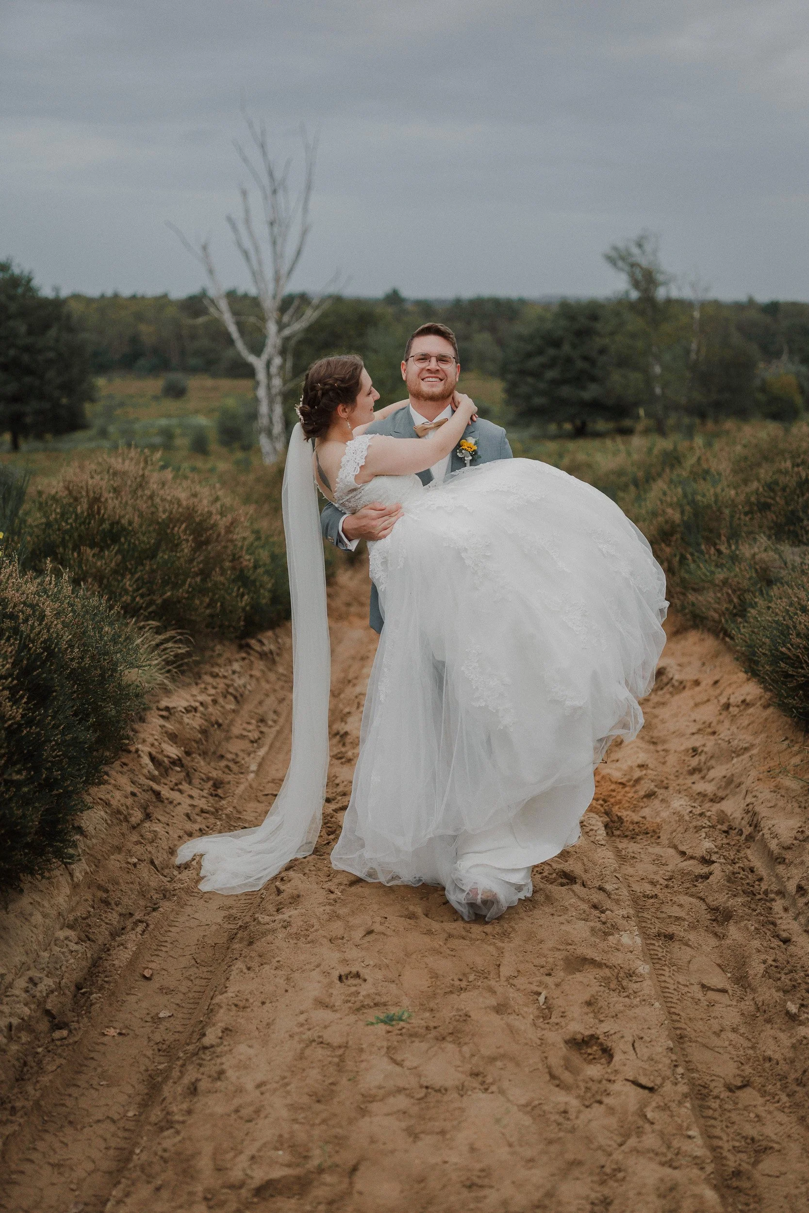 Ein Brautpaar in Hochzeitskleidung auf einem ländlichen Weg, der Bräutigam trägt einen Anzug und die Braut ein Hochzeitskleid, während der Bräutigam die Braut trägt. Im Hintergrund sind Bäume und ein bewölkter Himmel zu sehen.