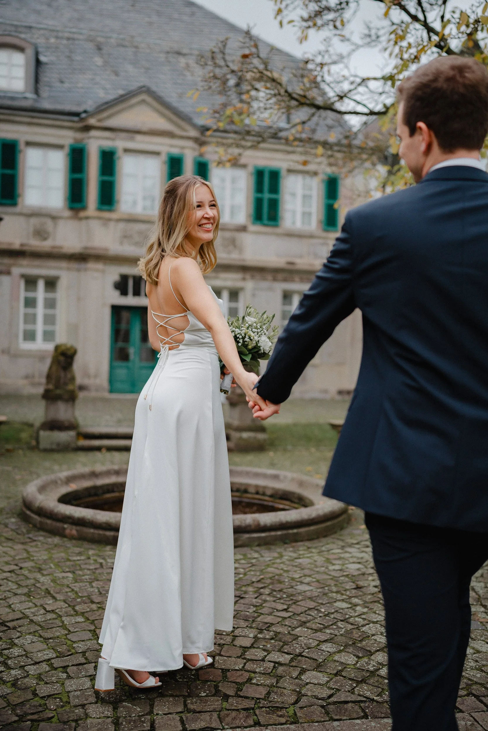 Ein glückliches Brautpaar hält sich an einem alten Brunnen auf einem gepflasterten Platz vor einem großen, historischen Gebäude mit grünen Fensterläden. Das Brautkleid ist lang und weiß mit Rückendetails, während der Bräutigam in einem dunklen Anzug 