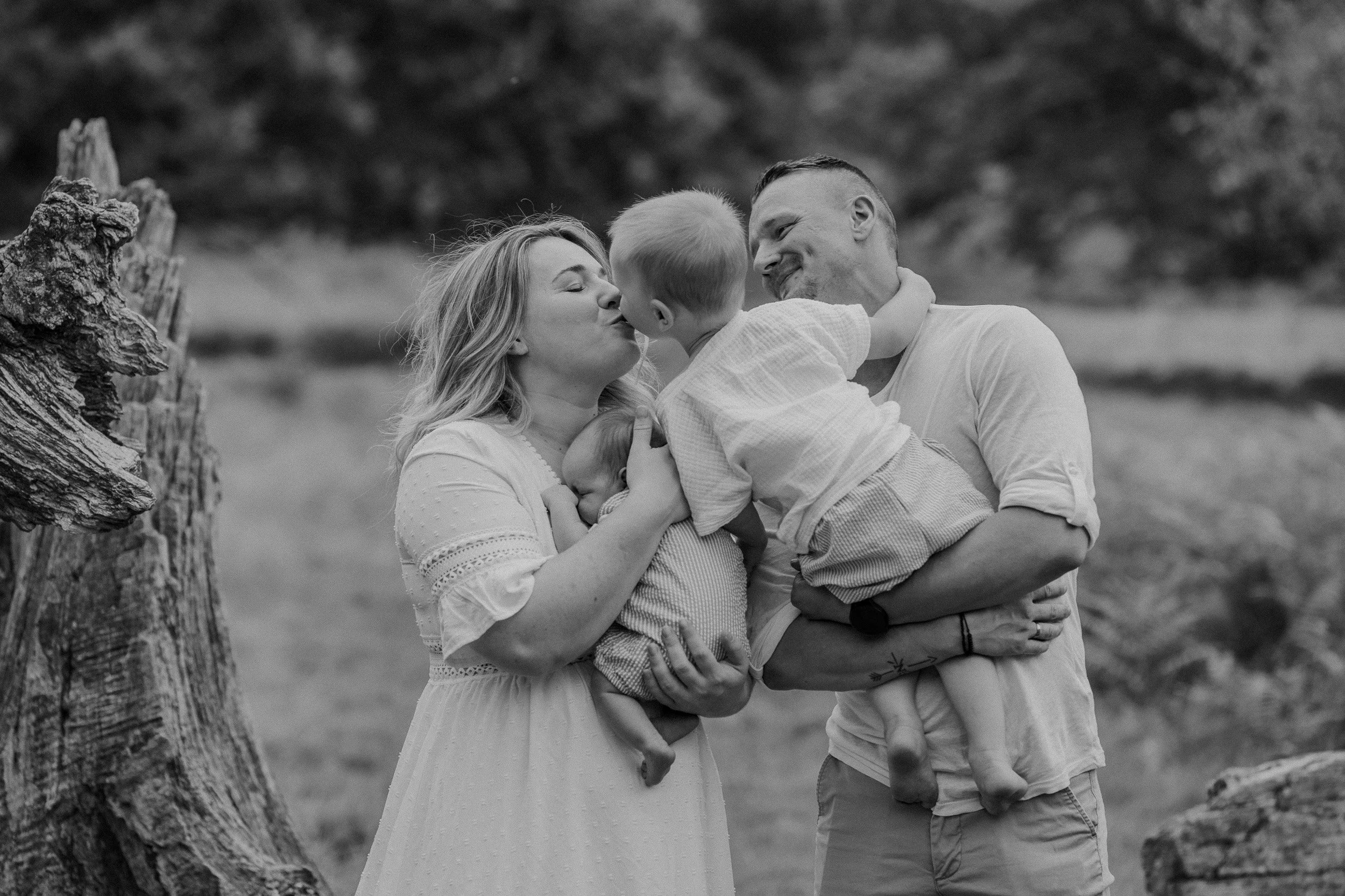 Familie mit Mutter, Vater, einem Baby und einem kleinen Kind im Freien, liebevoll umarmend und küssend, in natürlicher Umgebung, schwarz-weiß-Fotografie.