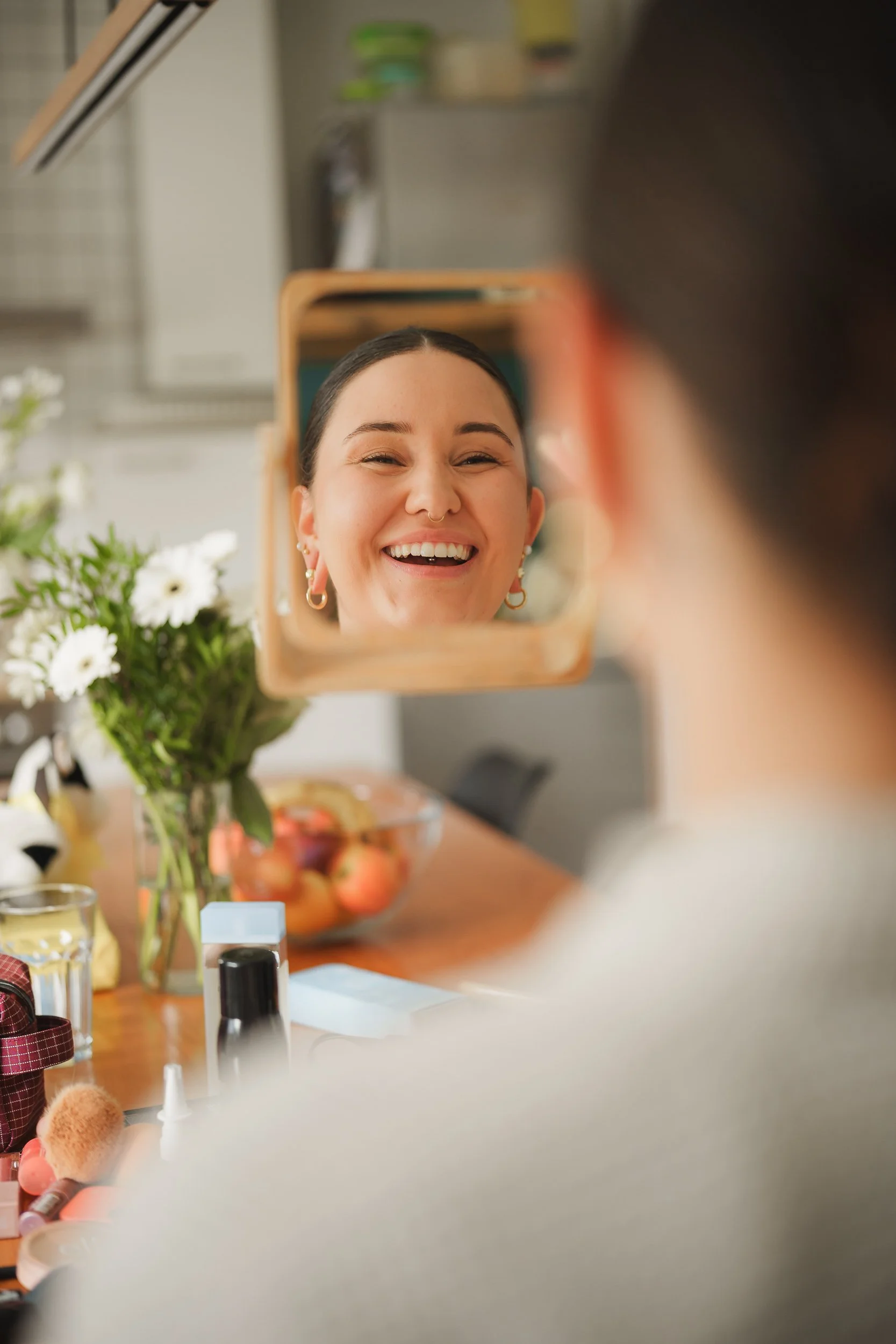 Frau lächelt beim Blick in einen kleinen Handspiegel, umgeben von Make-up-Produkten und einer Vase mit weißen Blumen auf einem Tisch.
