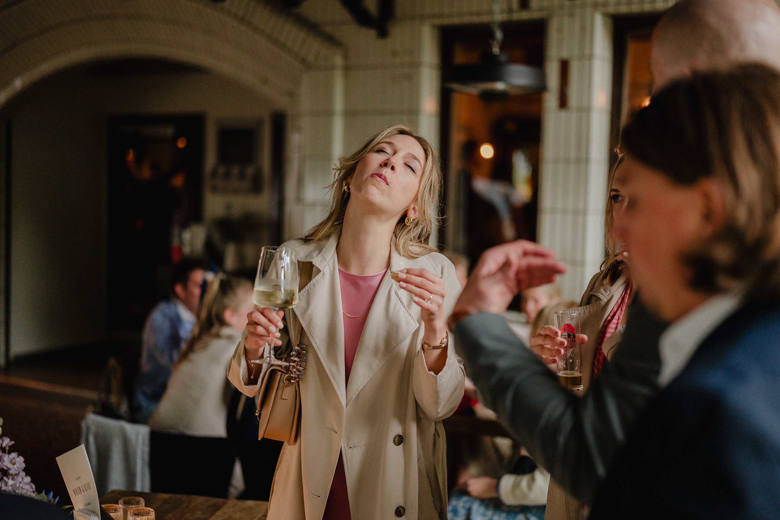 Frau in Trenchcoat mit Wein glas, die anscheinend genervt ist, während andere Leute im Hintergrund in einem Restaurant sitzen.