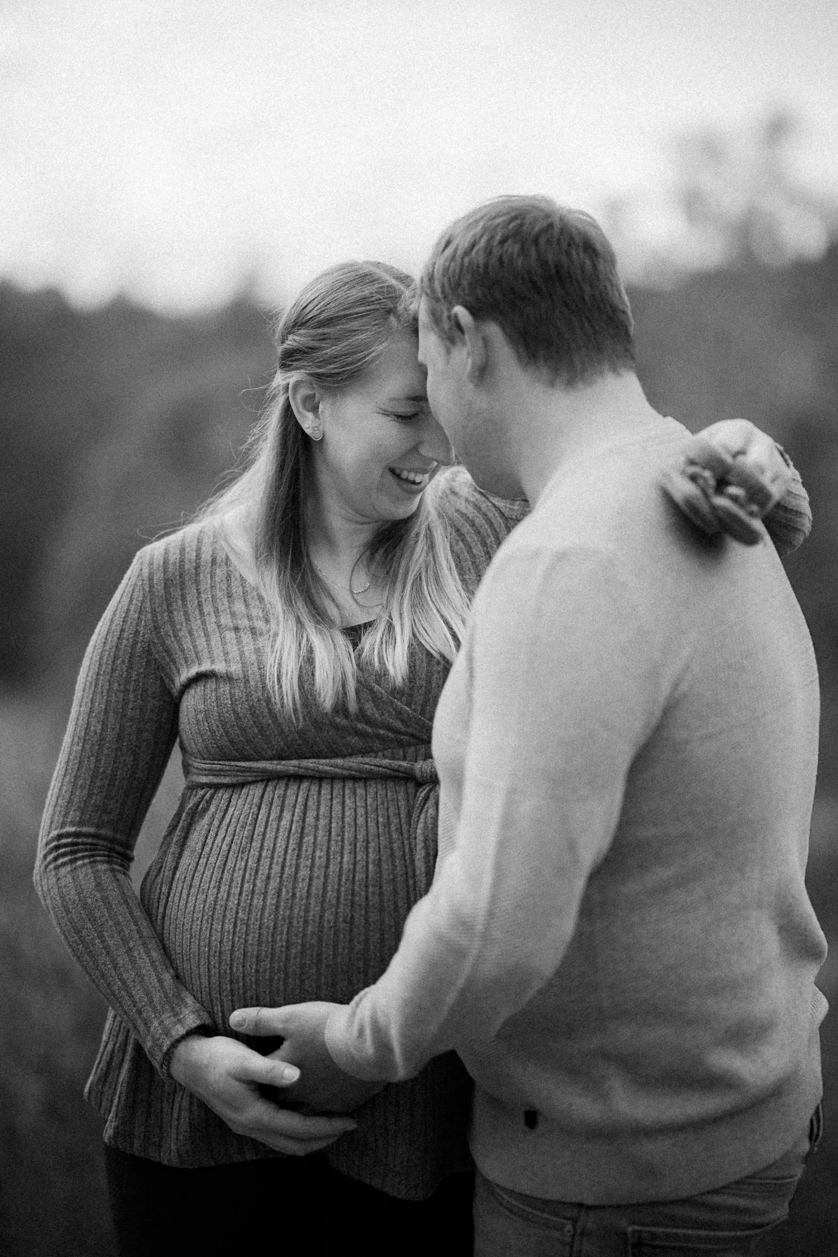 Ein schwangertes Paar, das sich liebevoll mit den Stirn aneinander und den Händen auf dem Babybauch berührt, im Freien bei Sonnenuntergang.