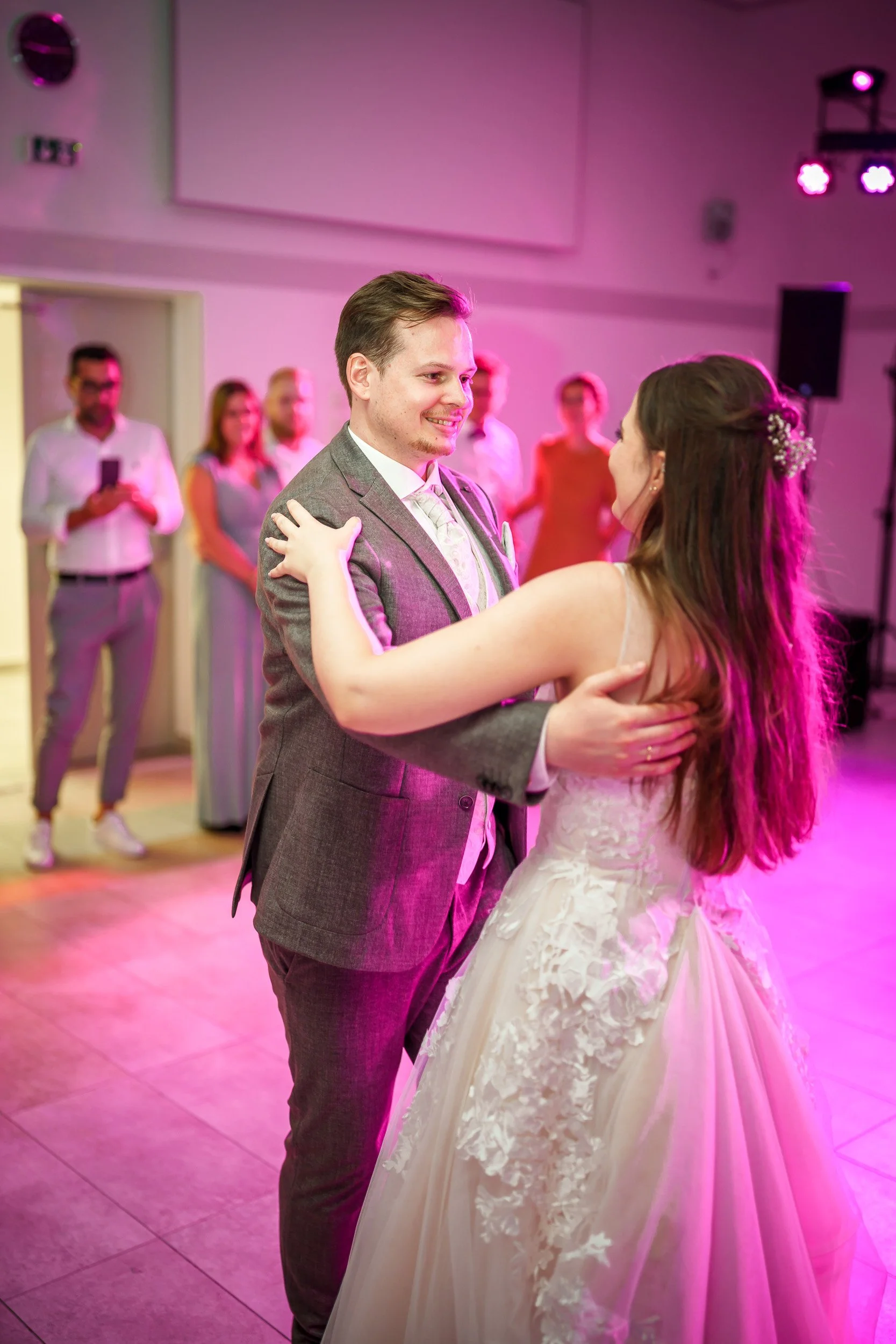 Ein Brautpaar tanzt auf einer Hochzeit, die Frau trägt ein weißes Brautkleid mit Spitze, der Mann trägt einen grauen Anzug.