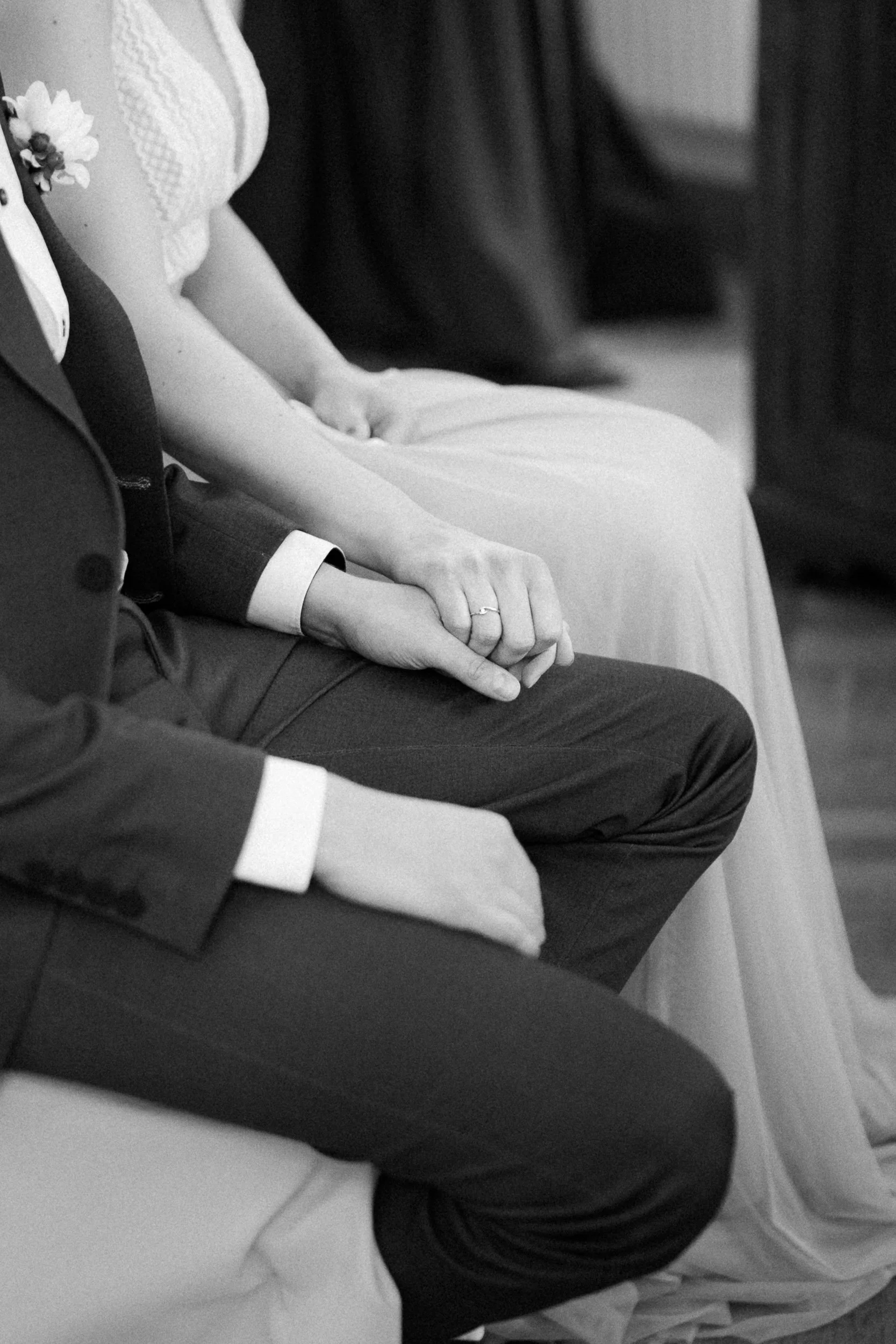 Nahaufnahme von einem Brautpaar bei einer Zeremonie, beide sitzen und halten sich an den Händen, die Frau trägt ein Hochzeitskleid, der Mann einen Anzug.