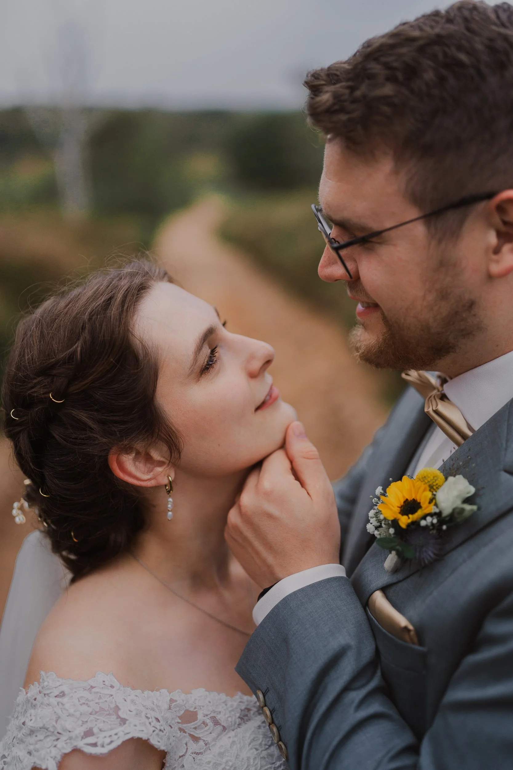 Ein Brautpaar schaut sich liebevoll in die Augen. Der Mann hält die Frau sanft am Kinn, während sie glücklich lächelt. Sie trägt eine Hochzeitskleidung und steht im Freien auf einem Weg.