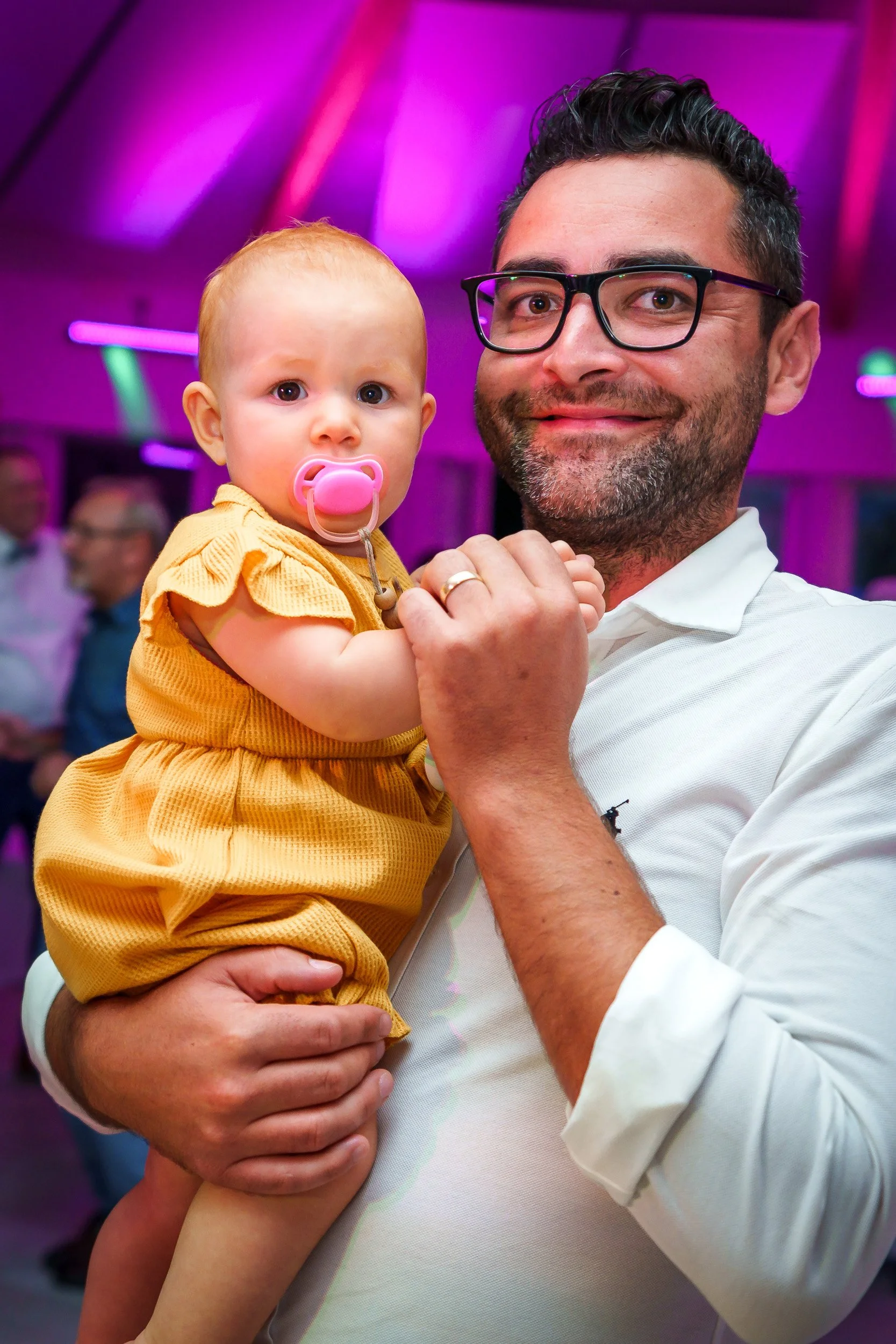 Ein Mann hält ein Baby mit einem Schnuller in einem gelben Kleid in einer bunten, diskothekenähnlichen Umgebung.