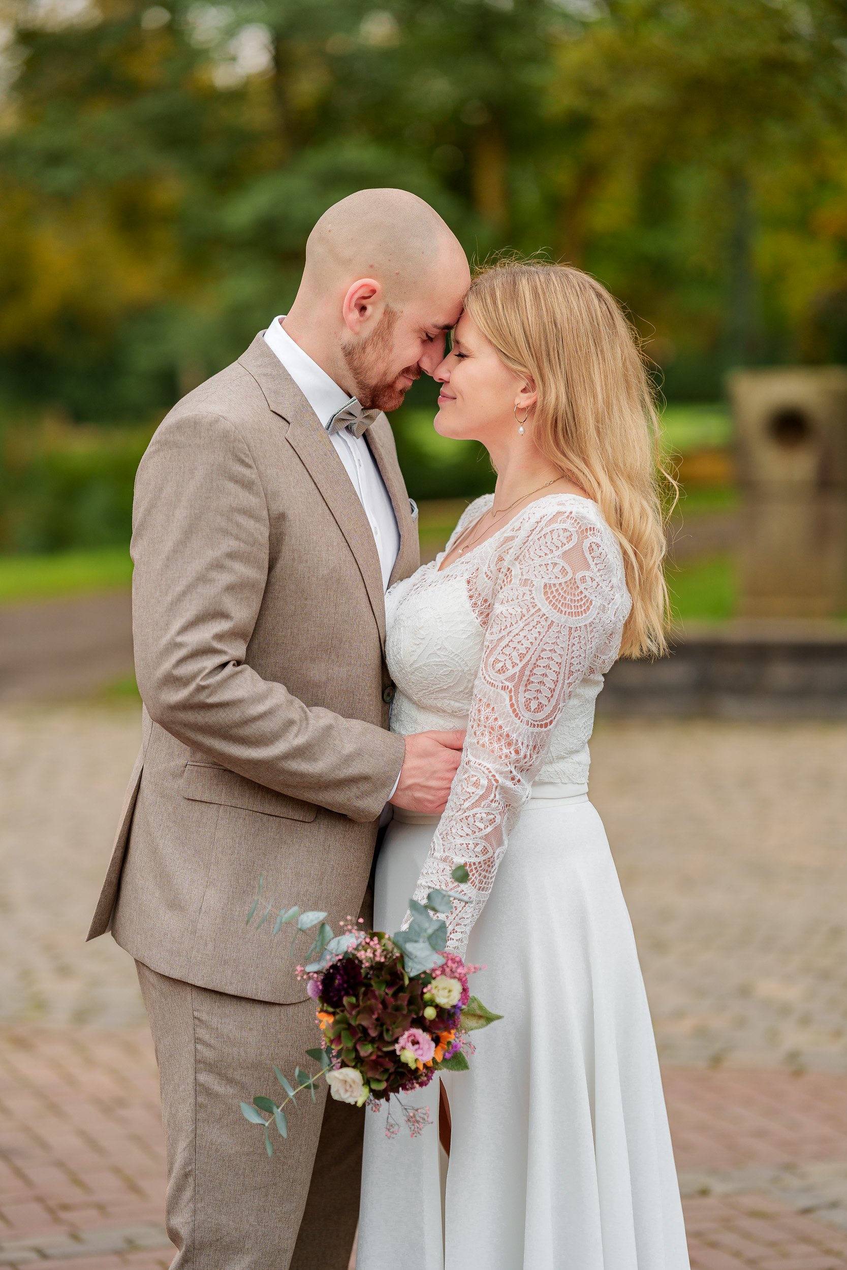 Ein Brautpaar steht sich gegenüber, beide mit geschlossenen Augen, in einem Park bei Sonnenuntergang, die Stirn berührend, die Braut hält einen bunten Blumenstrauß.
