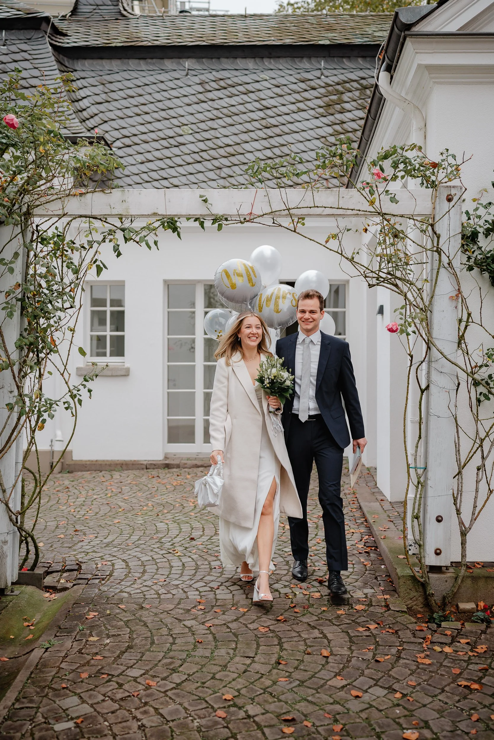 Ein glückliches Brautpaar läuft zusammen unter einem weißen Torbogen mit rosafarbenen Rosen und Heu, das auf den Bögen wächst. Der Braut trägt ein weißes Kleid mit einem hohen Schlitz und einen weißen Mantel, während der Bräutigam einen dunklen Anzug