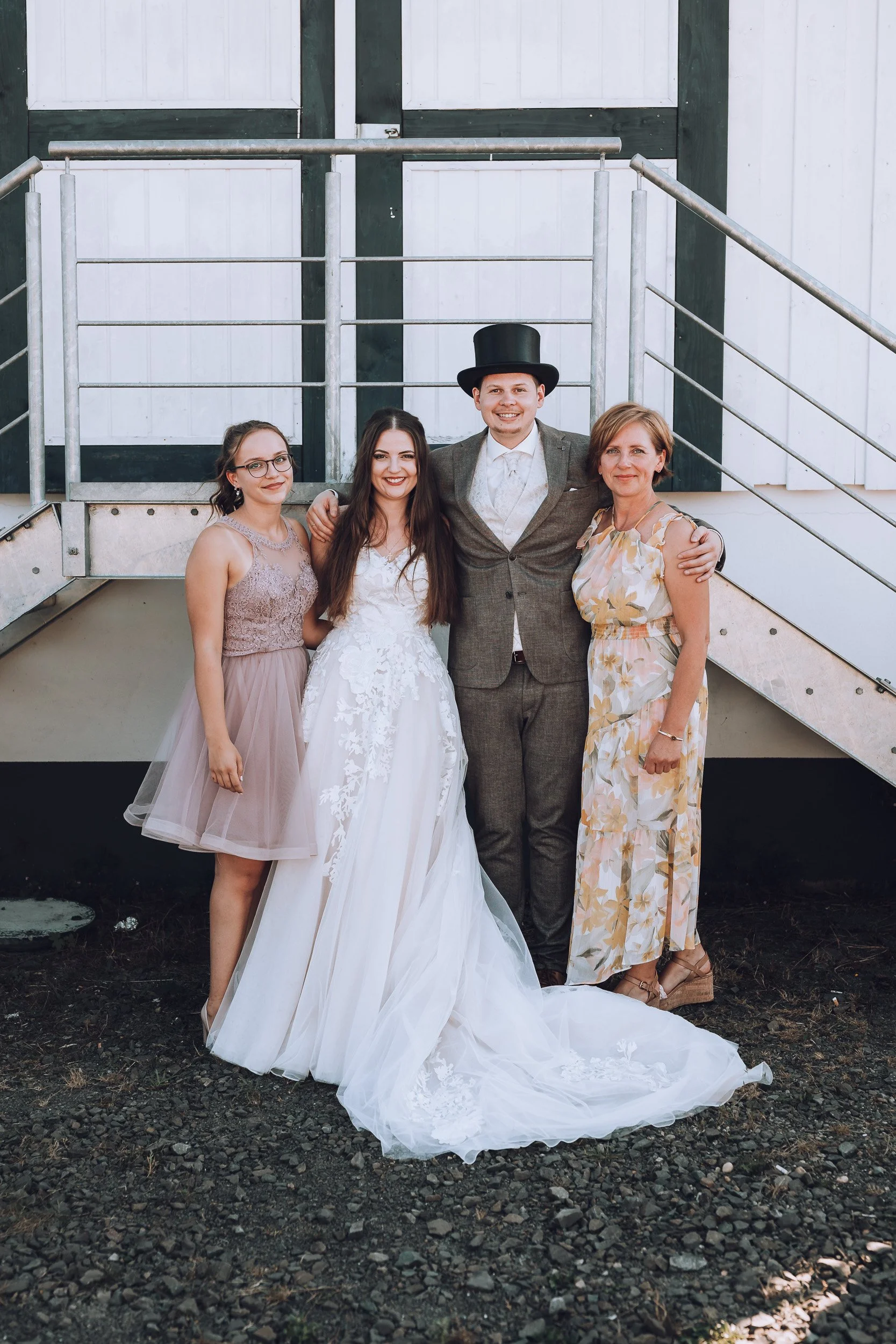 Gruppe von fünf Menschen bei einer Hochzeit, darunter eine Braut, ein Bräutigam mit Zylinderhut und drei Frauen, die vor einer scheunenähnlichen Kulisse stehen.