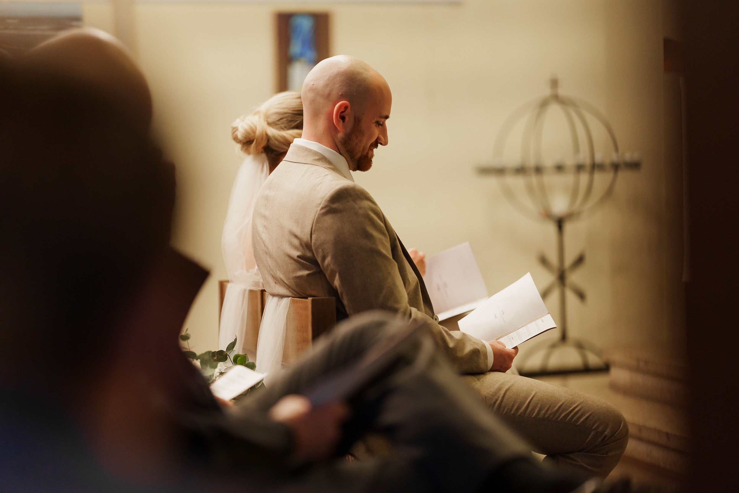 Ein Mann und eine Frau sitzen in einer Zeremonie, lesen aus Heften oder Programmen, vermutlich bei einer Hochzeit, mit Kerzen und Dekoration im Hintergrund.