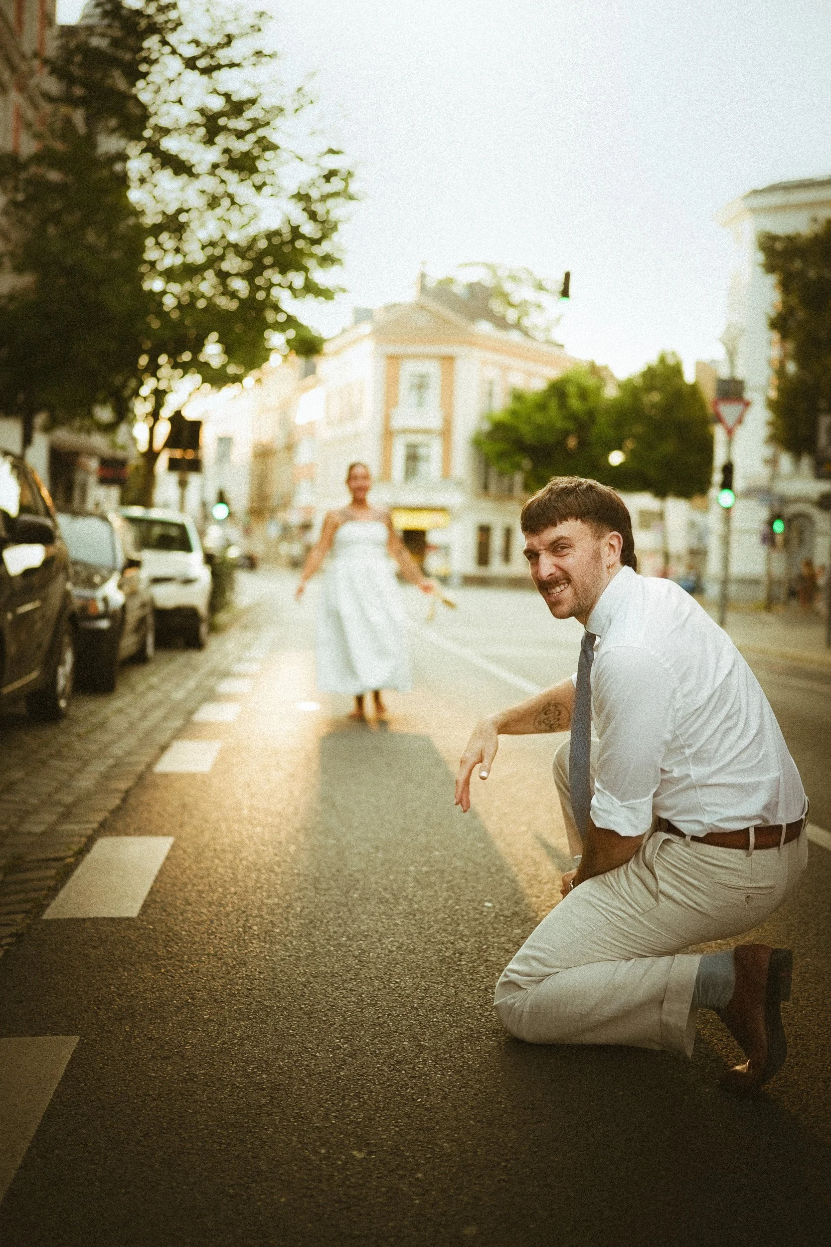 Ein Mann kniet auf einer Straße, trägt weiße Hemd und beige Hosen, lächelt in die Kamera, im Hintergrund eine Frau in einem weißen Kleid, auf einer Stadtstraße bei Sonnenuntergang.