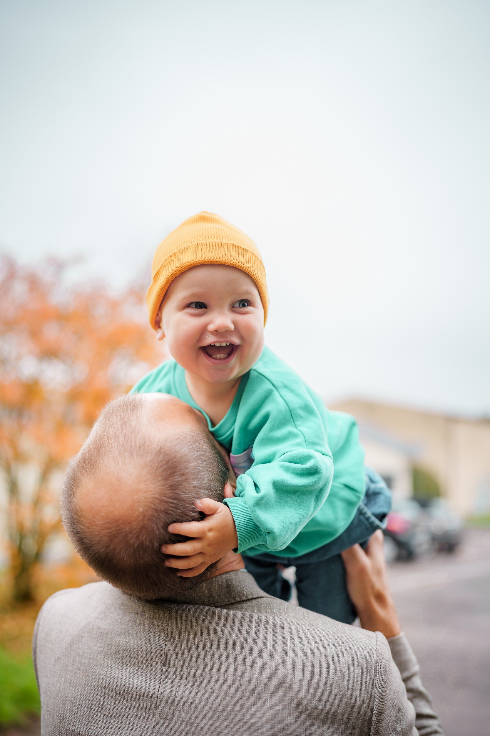 Lächelndes Kleinkind auf den Schultern eines Mannes mit Baum im Herbst im Hintergrund.