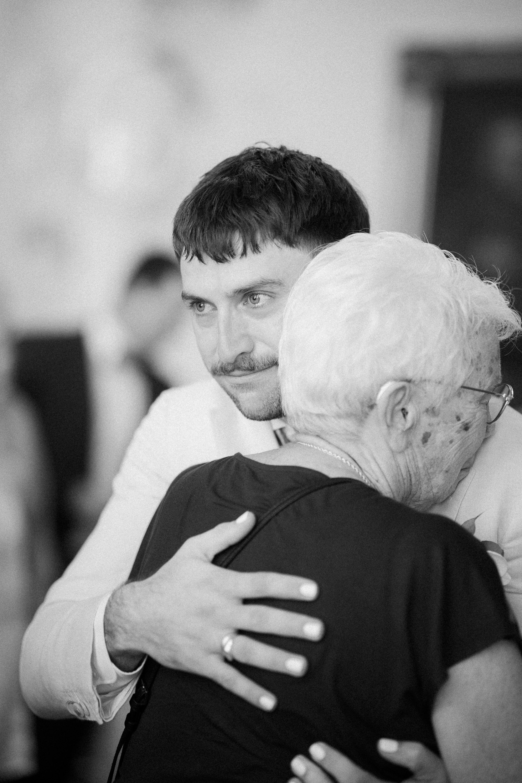 Ein junger Mann umarmt eine ältere Frau bei einem Tanz oder einer Feier, im Hintergrund sind weitere Personen sichtbar.