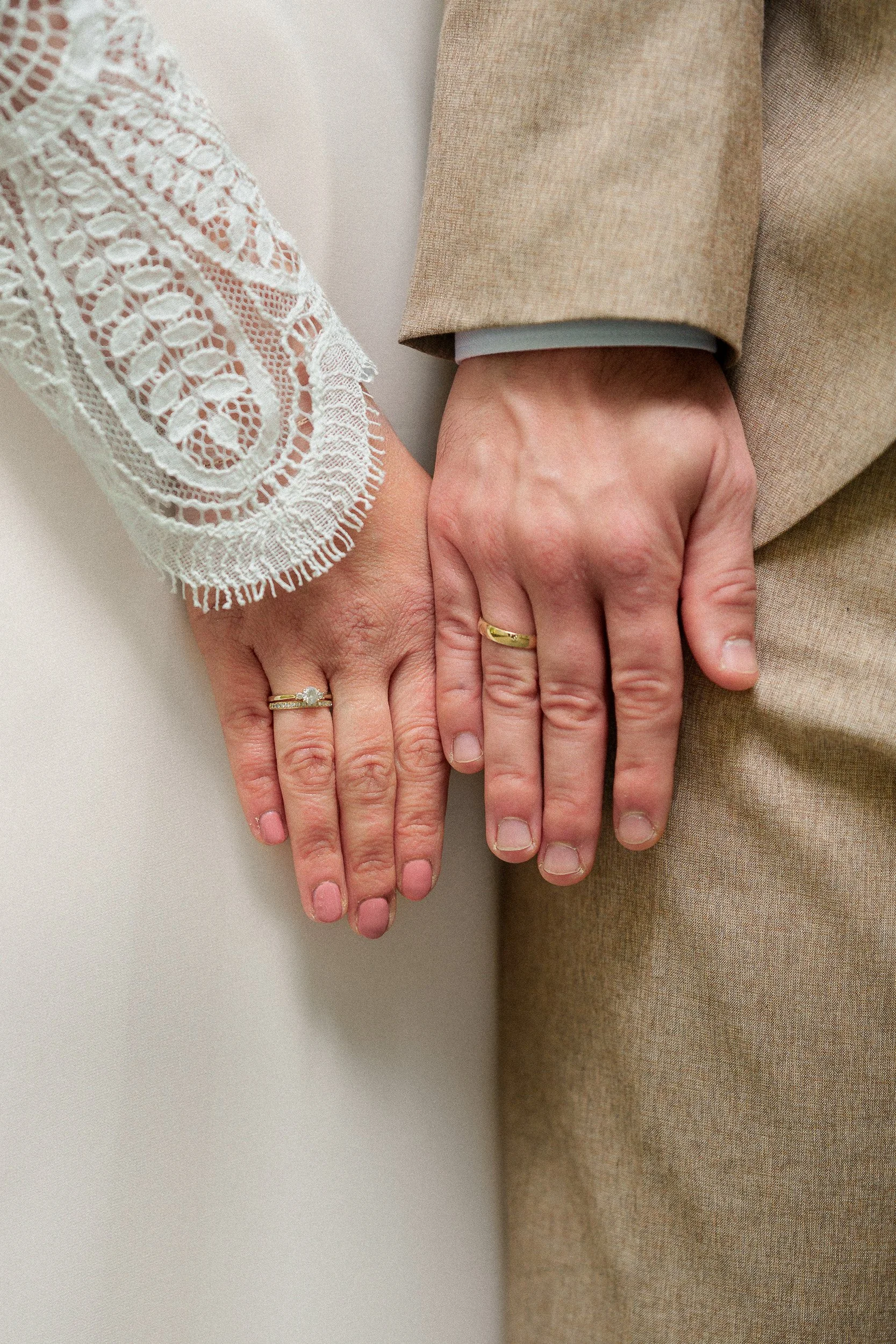 Nahaufnahme der Hände eines Paares, die Hand in Hand liegen, mit Eheringen, eine Person trägt eine beige Jacke und eine andere Person trägt eine weiße Bluse mit Spitze.