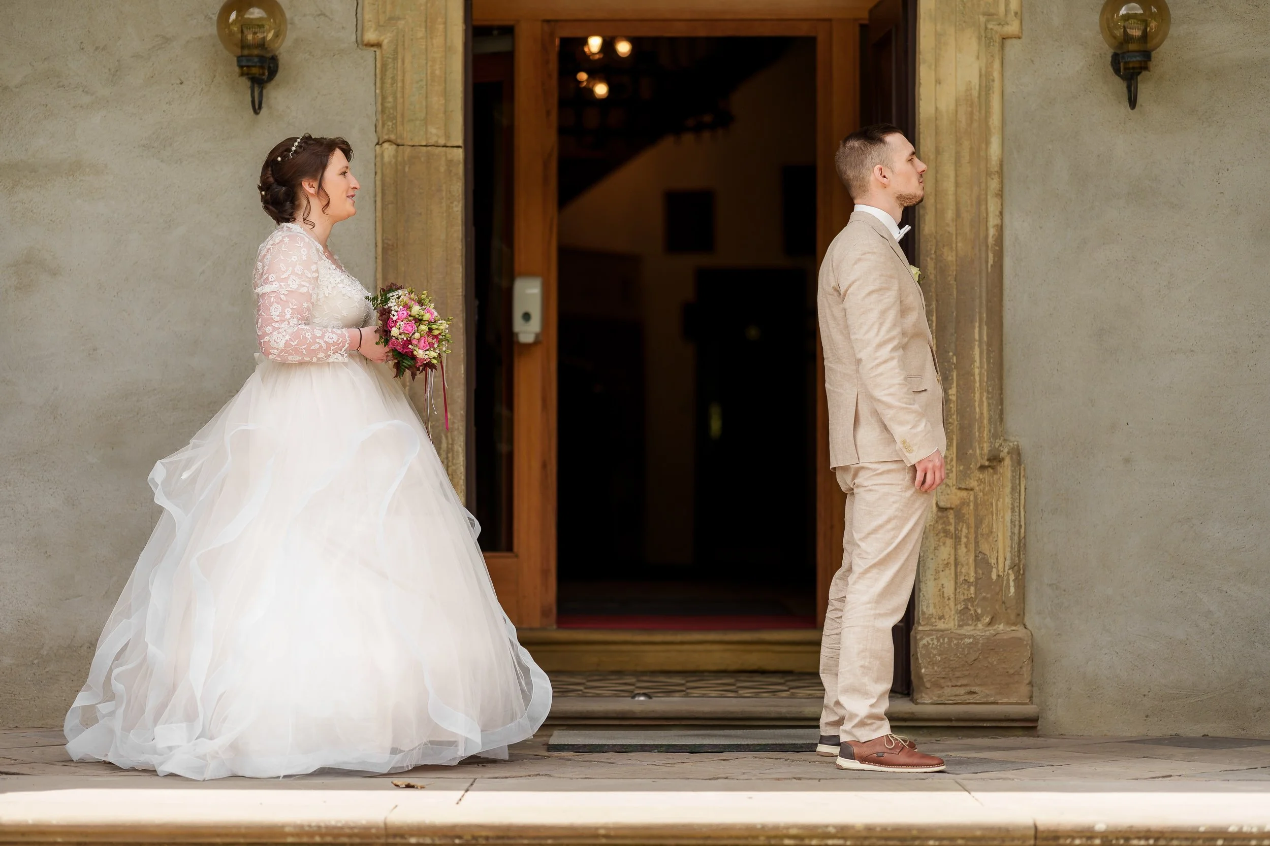 Braut und Bräutigam stehen getrennt vor der Tür eines Hauses, mit Abstand zueinander, in Hochzeitskleidung.