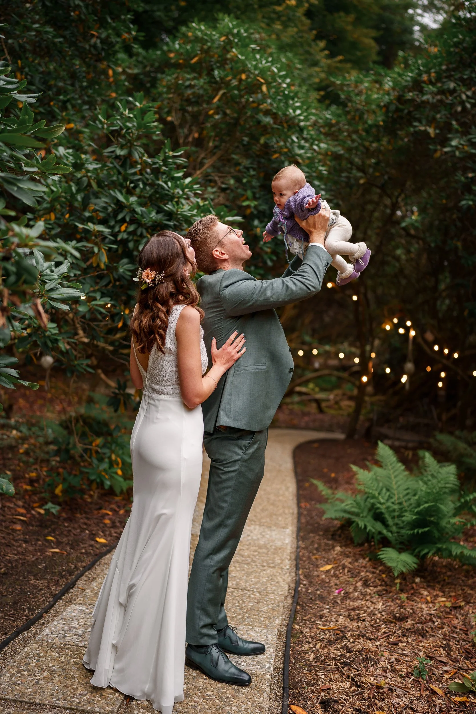 Ein frisch vermähltes Paar, ein Mann und eine Frau, steht auf einem gepflasterten Weg in einem Garten mit grünen Pflanzen und Bäumen, während der Mann ein Baby hoch in die Luft hebt. Es ist Abenddämmerung mit warmem Licht und Lichterketten im Hinterg