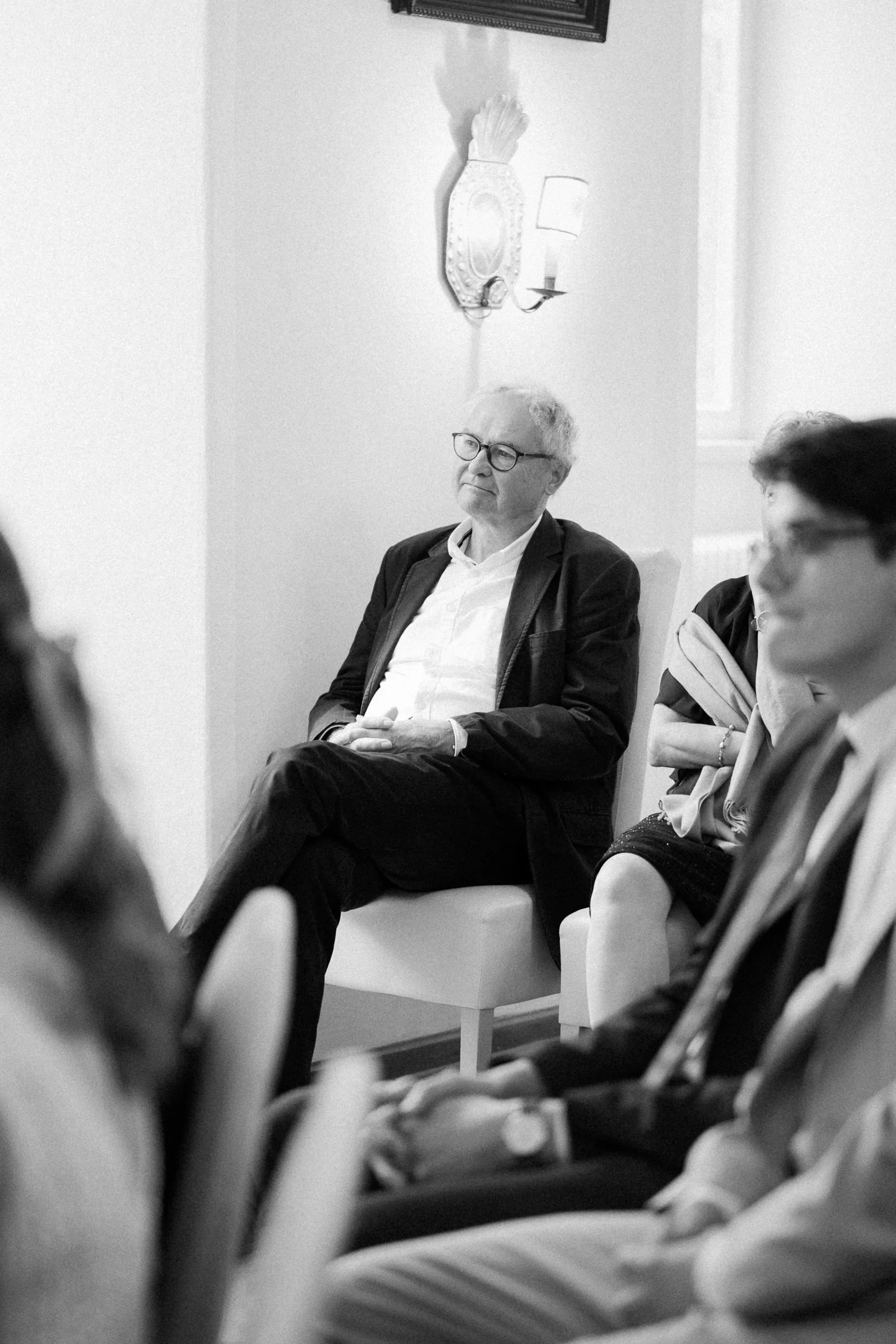 Schwarz-weiß Foto eines Mannes mit Brille, sitzend in einem Raum, umgeben von anderen Personen, mit einer Wandlampe und Dekoration an der Wand im Hintergrund.