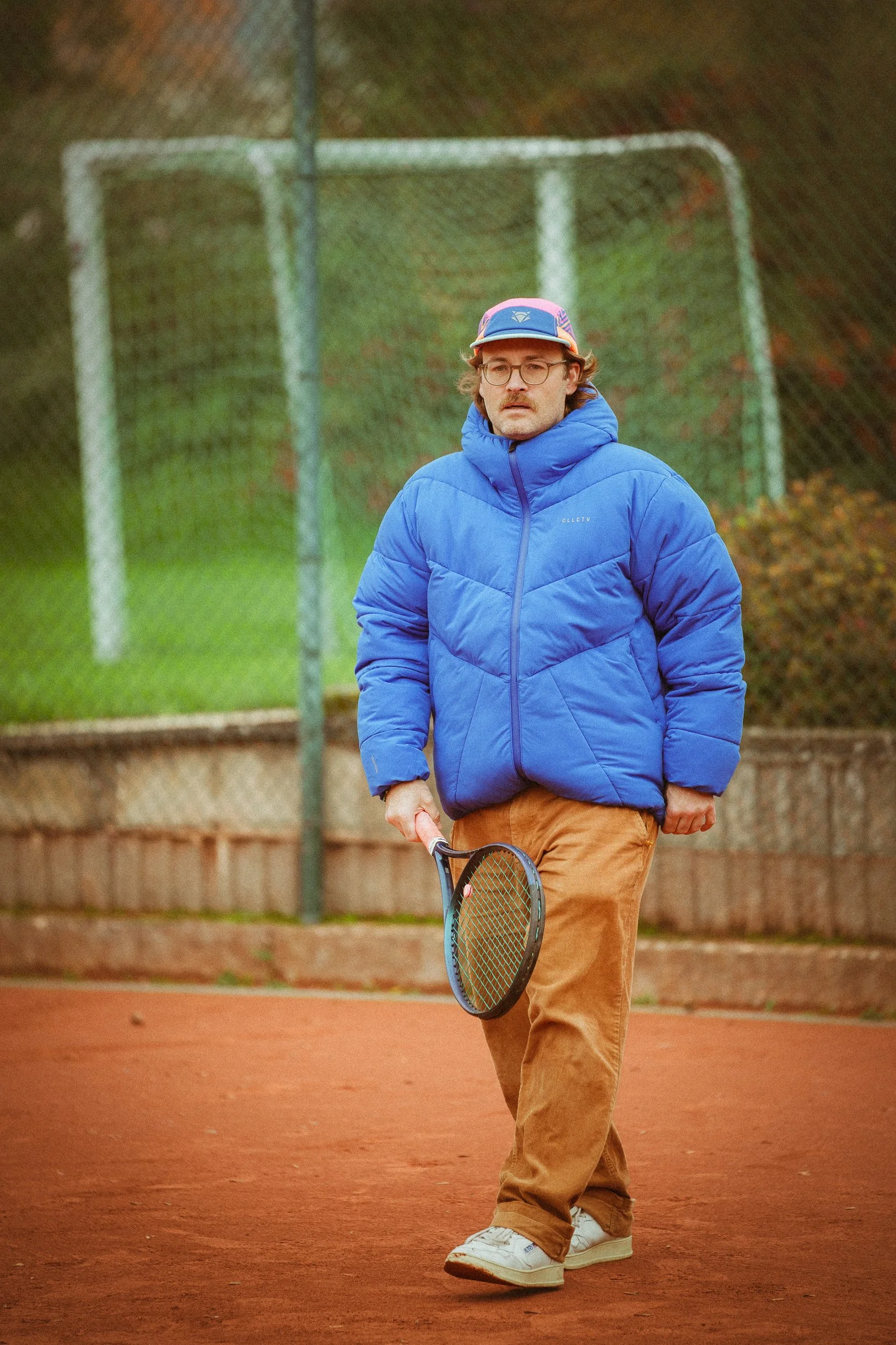 Mann auf einem Tennisplatz, trägt eine blaue Jacke, Brille und einen bunten Hut, hält einen Tennisschläger in der Hand, im Hintergrund ein Tennisnetz.