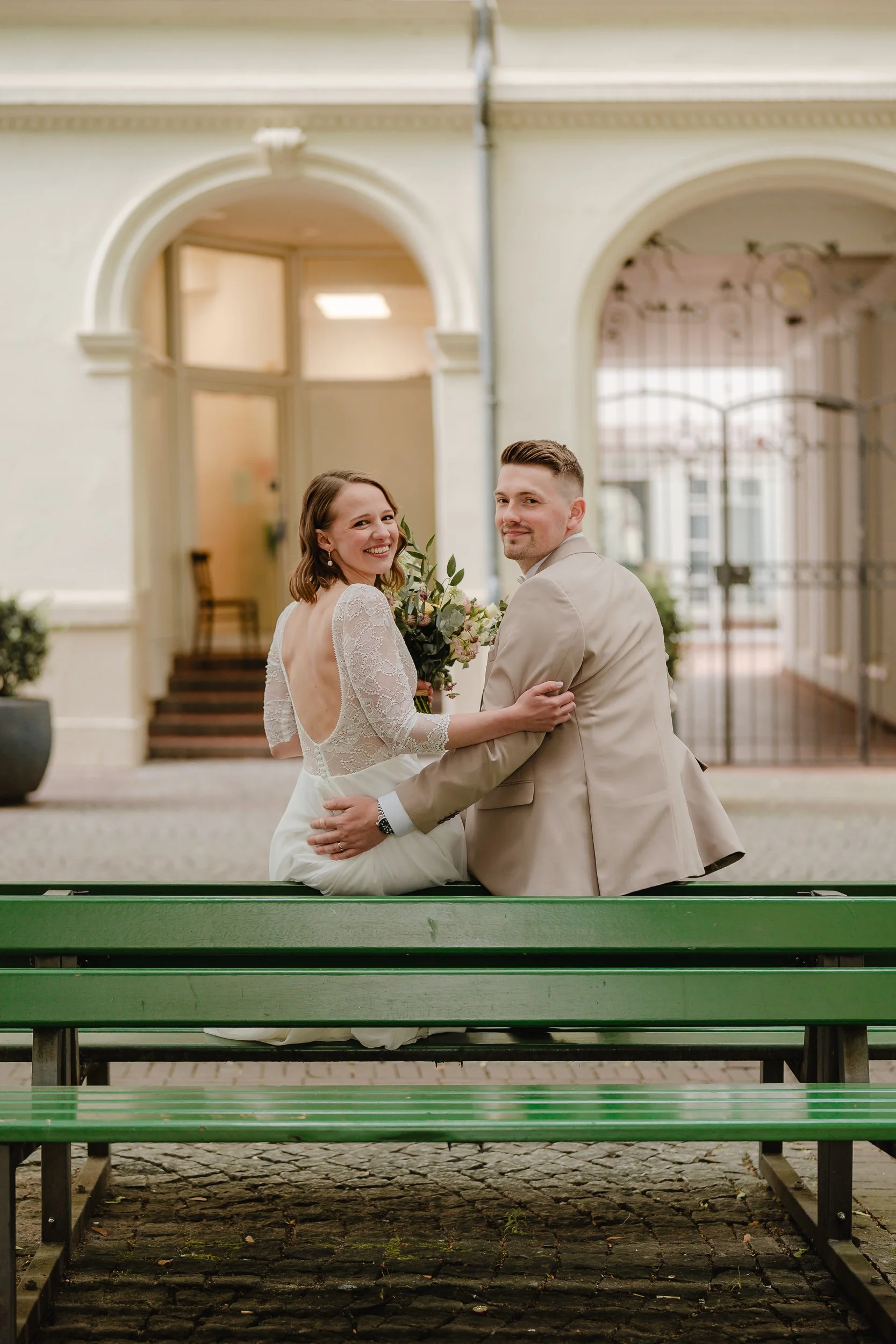 Ein frisch verheiratetes Paar sitzt auf einer Parkbank vor einem historischen Gebäude, lächelt, die Frau hält einen Blumenstrauß, die Szene ist hell und freundlich.