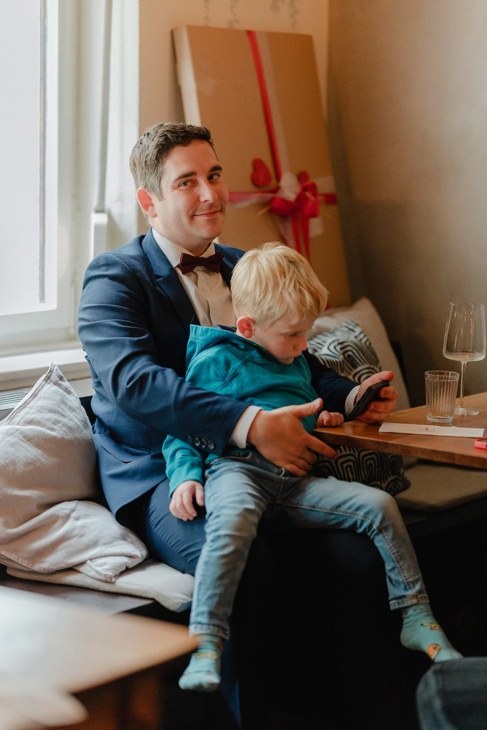 Ein Mann im Anzug und Krawatte sitzt auf einem Sofa neben einem jungen Jungen, der auf seinem Schoß sitzt und ein Smartphone hält, während sie zusammen an einem Tisch bei einem festlichen Anlass sitzen.