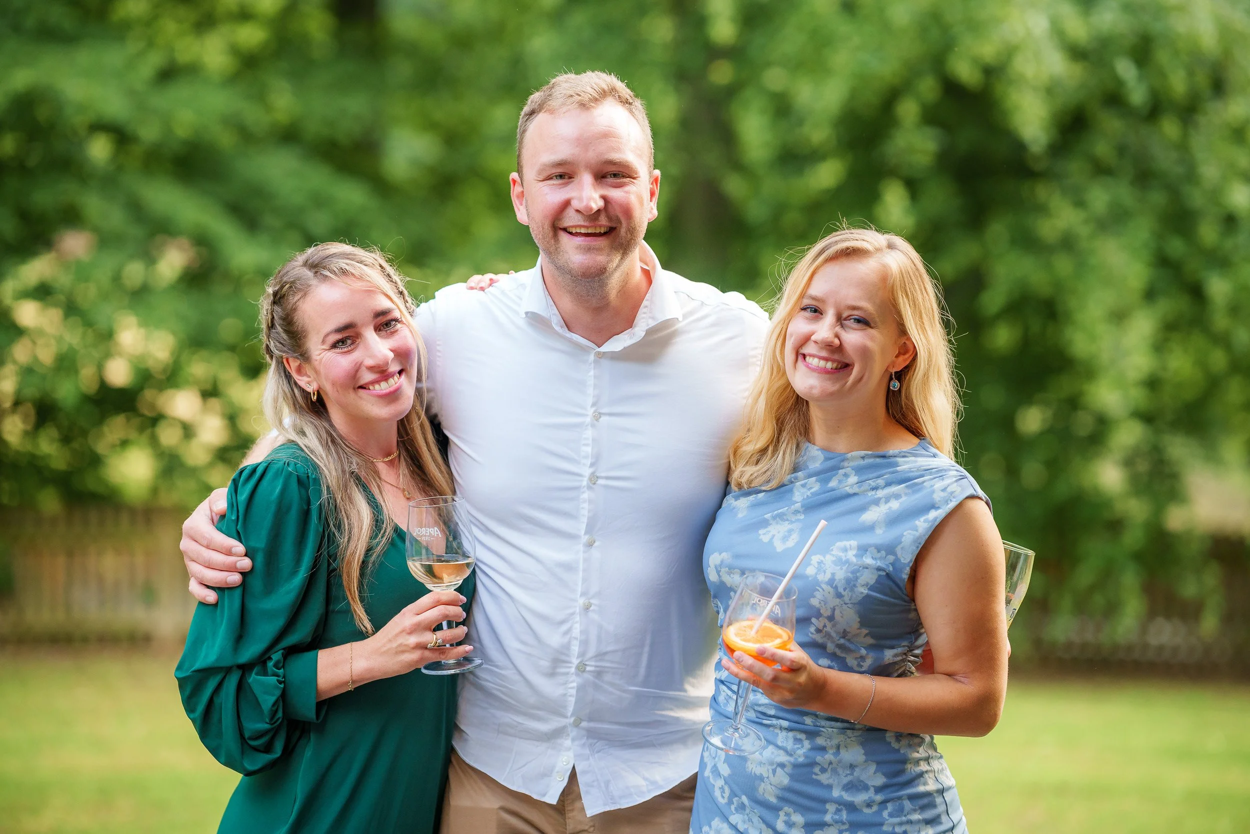 Drei Menschen, zwei Frauen und ein Mann, posieren im Freien mit Getränken bei einer Feier. Der Mann trägt ein weißes Hemd, die Frauen tragen elegante Kleider, und alle lachen.