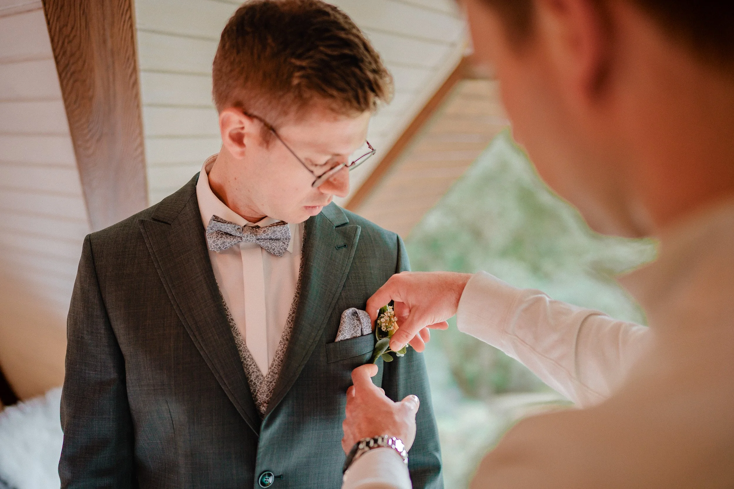 Eine Person steckt einem jungen Mann einen Blumenanstecker in die Brusttasche seines Anzugs, während er nach unten schaut.