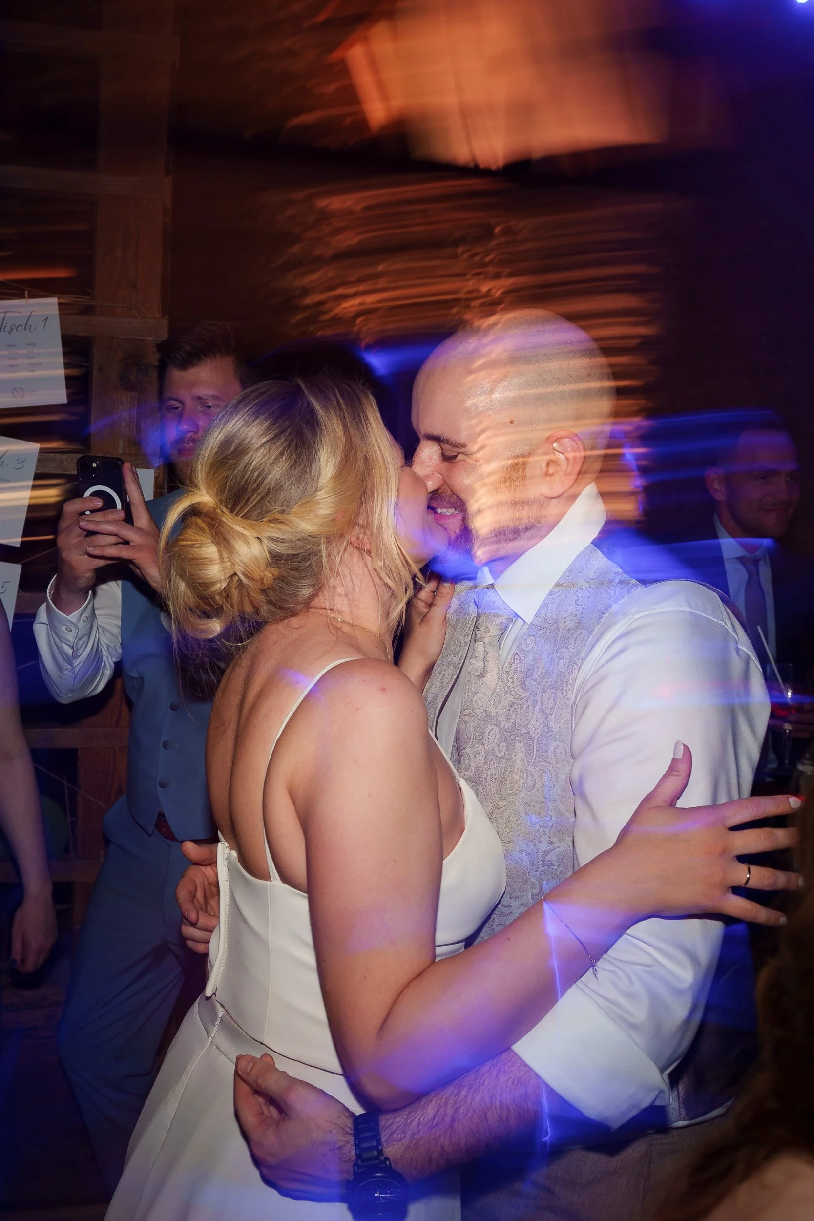 Ein Paar bei einer Hochzeitsfeier umarmt sich und küsst sich, andere Gäste im Hintergrund, bunte Lichteffekte.