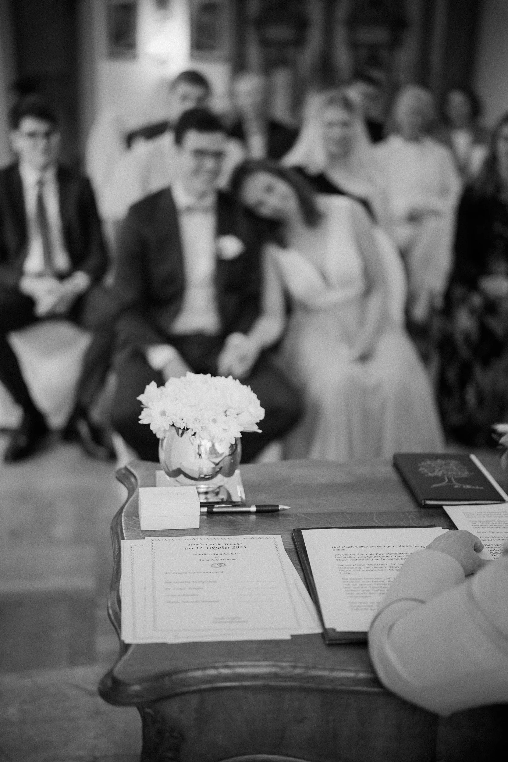 Ein festlicher Anlass, vermutlich eine Hochzeit, mit Schreibmaterialien, Blumen und einem Gast im Vordergrund, im Hintergrund eine Gruppe von Menschen in formeller Kleidung, die auf einem Sofa sitzen.