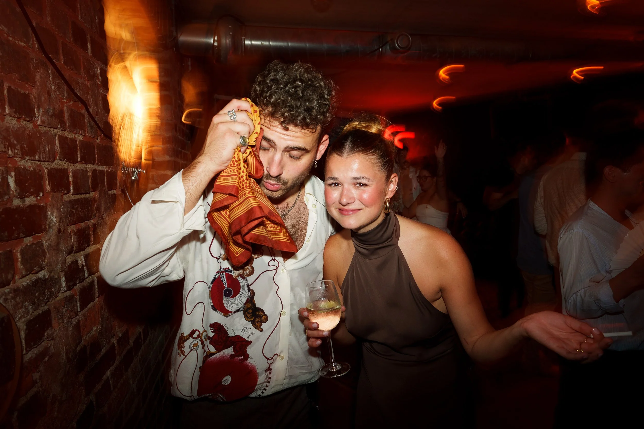 Zwei junge Menschen auf einer Party, die in einem dunklen Raum mit roten und orangefarbenen Lichtern stehen. Der Mann trägt ein weißes Hemd mit bunten Mustern und hat einen Schal um den Hals, während die Frau ein braunes Kleid trägt und ein Glas Wein