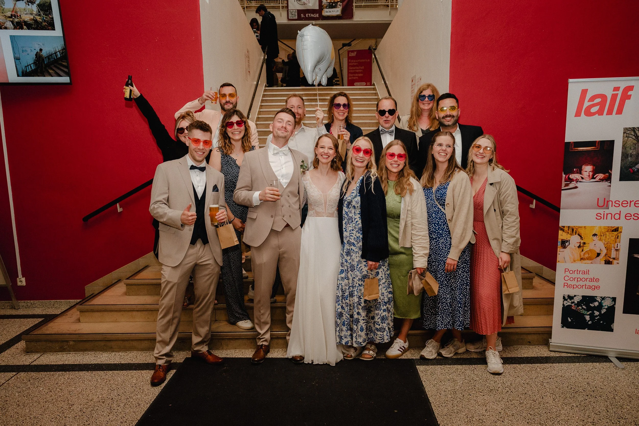 Gruppe von Menschen bei einer Feier, einige tragen Anzüge und Kleider, alle tragen Sonnenbrillen und lachen, im Hintergrund eine rote Wand und eine Treppe.