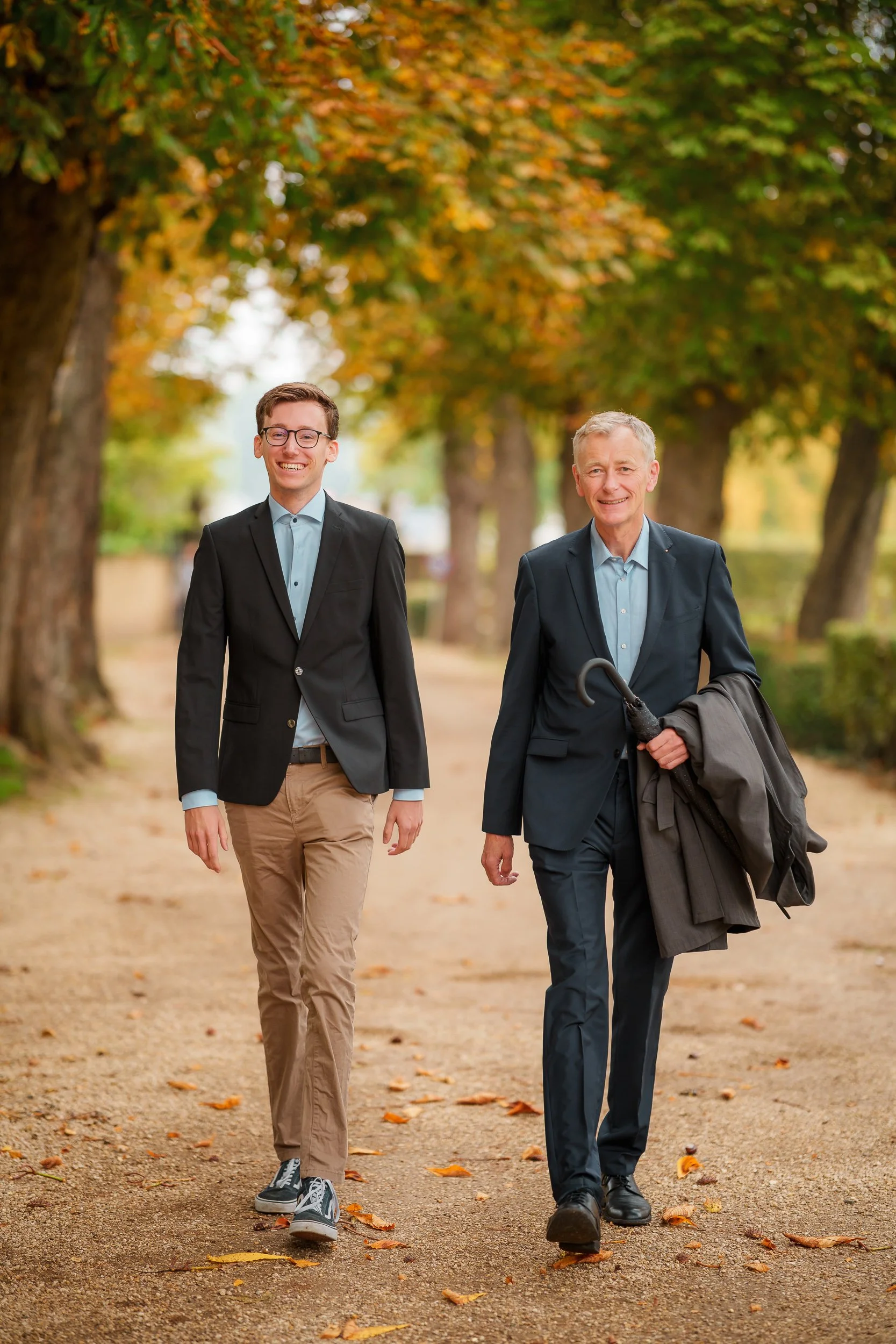 Zwei Männer in Anzügen spazieren durch einen herbstlichen Park mit fallen Laub und bunten Bäumen.