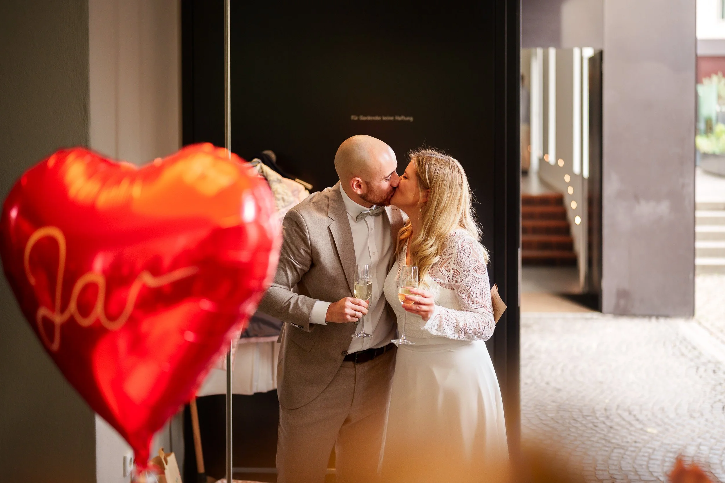 Ein Paar bei einer Hochzeit küsst sich, beide hält Sektgläser in der Hand, im Vordergrund ist ein rotes Herzballon mit der Aufschrift 'Love' sichtbar.