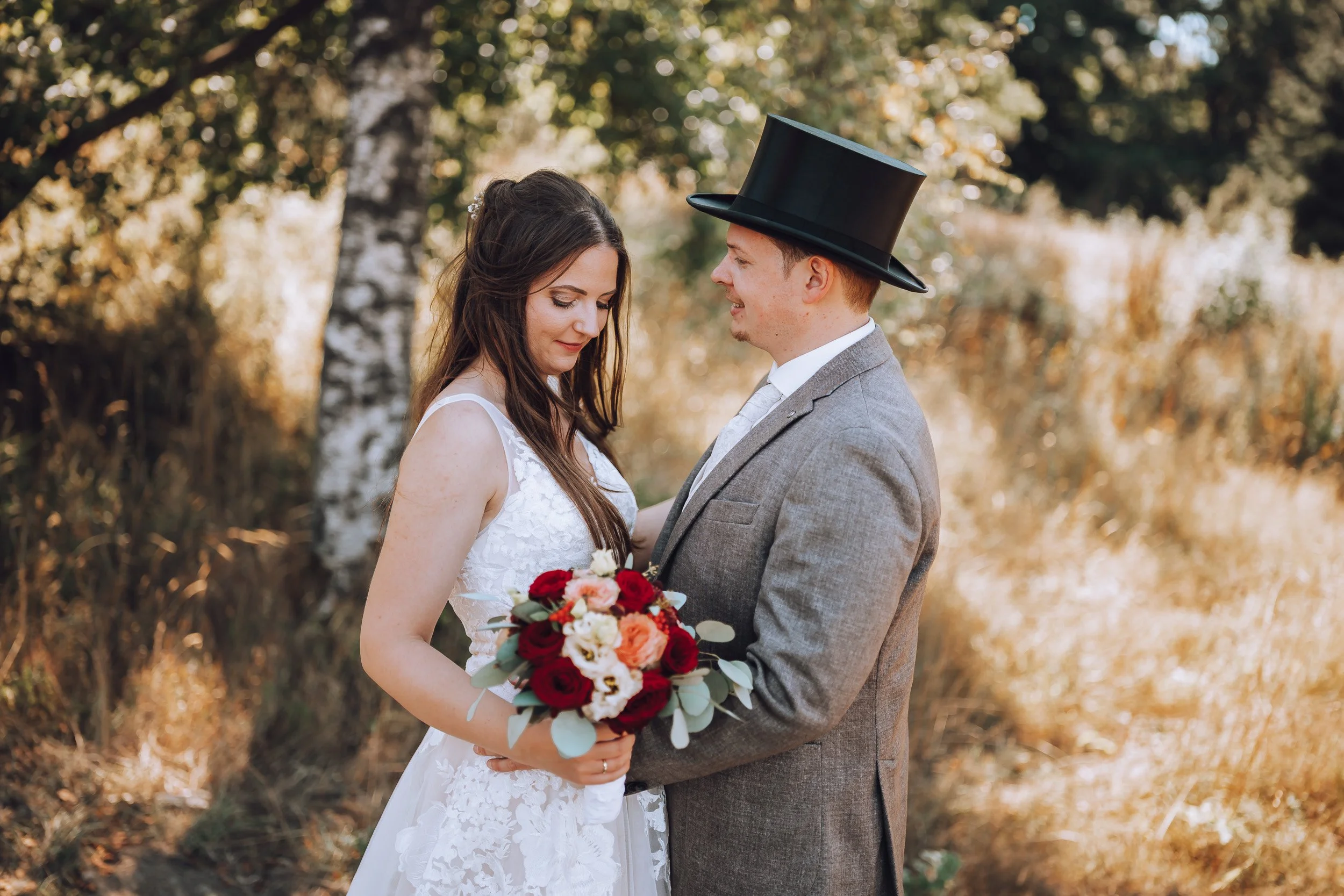 Ein Brautpaar, die Braut in einem weißen Kleid und der Bräutigam in einem Anzug mit Zylinder, stehen sich im Freien gegenüber, die Braut hält einen Blumenstrauß, im Hintergrund Bäume mit herbstlicher Laubfärbung.