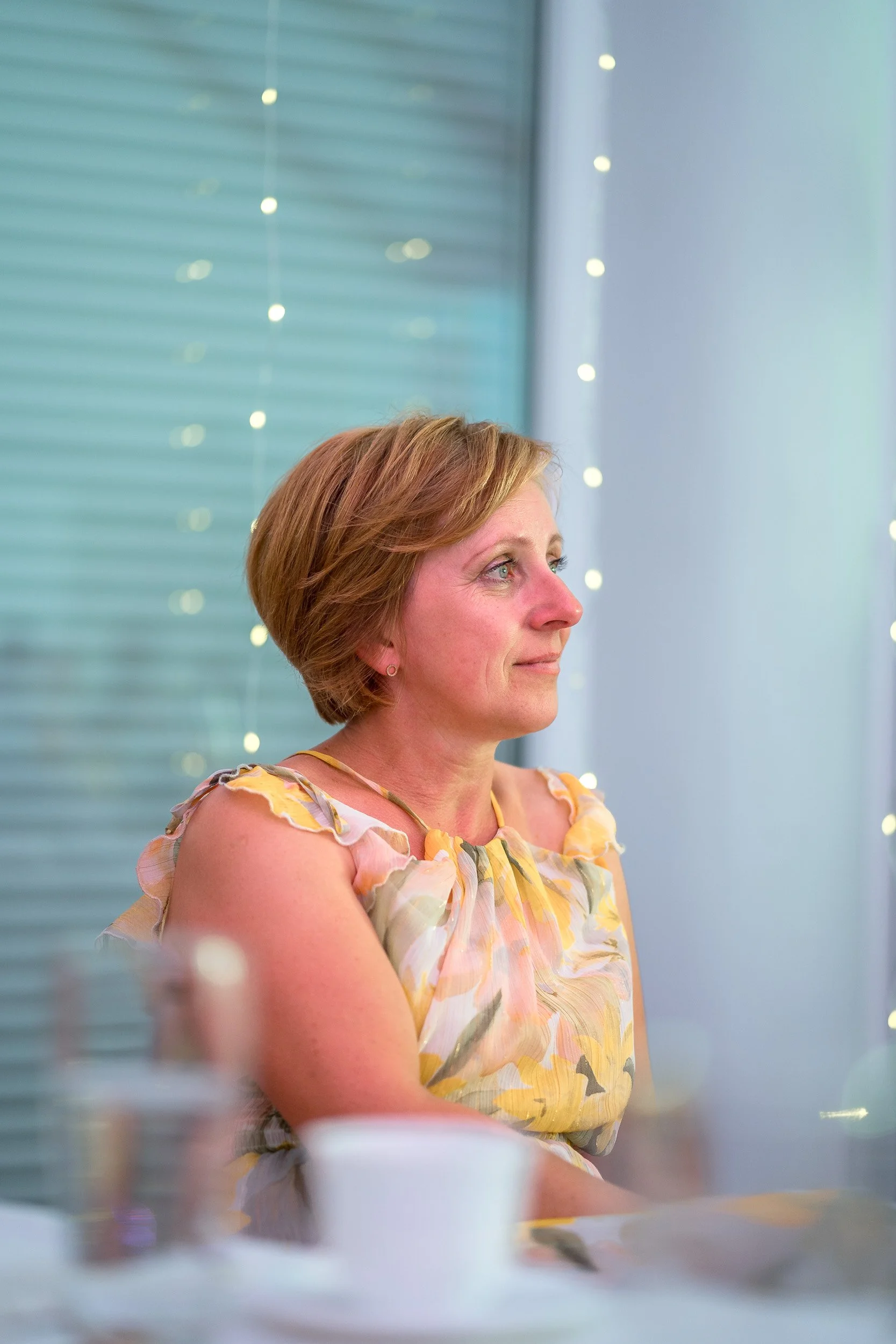 Frau mit kurzen braunen Haaren in einem bunt gemusterten Sommerkleid, sitzt auf einem Stuhl in einem Raum mit blauen Vorhängen und Lichterketten im Hintergrund.