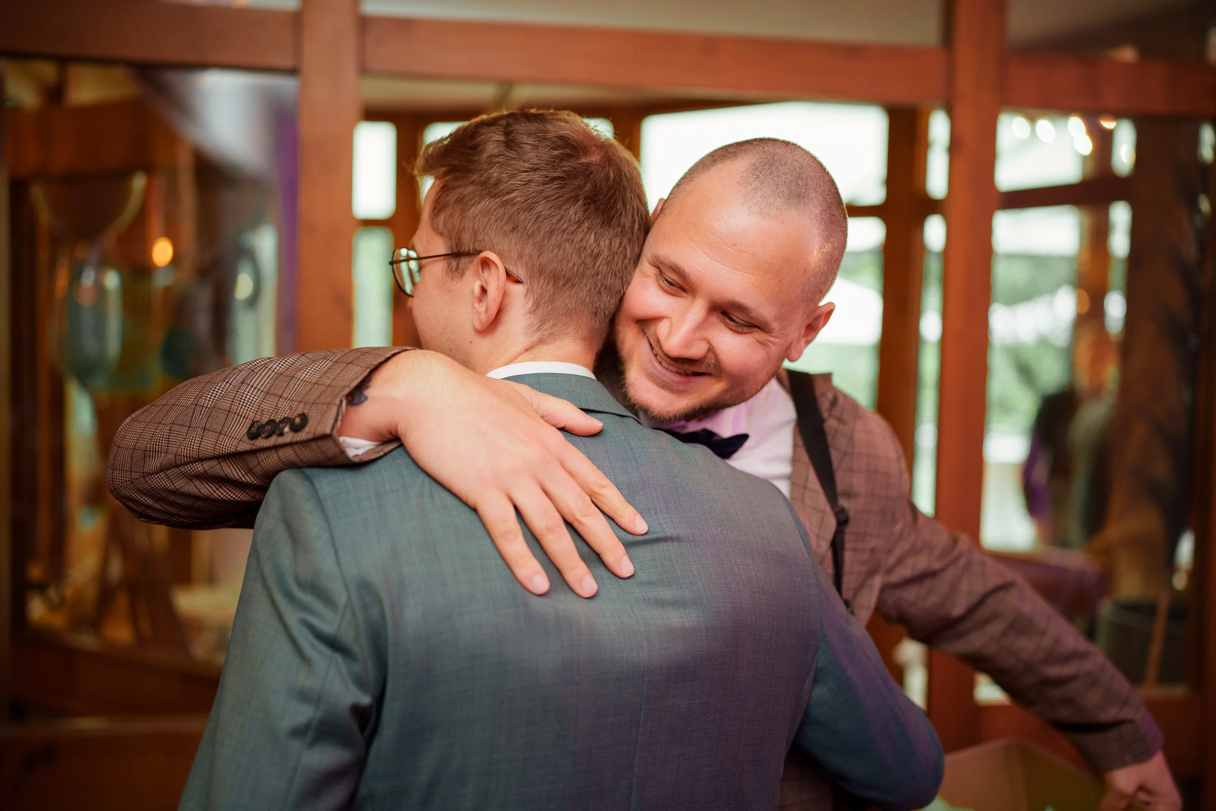 Zwei Männer umarmen sich herzlich bei einer Feier, im Hintergrund Holzmöbel und große Fenster.