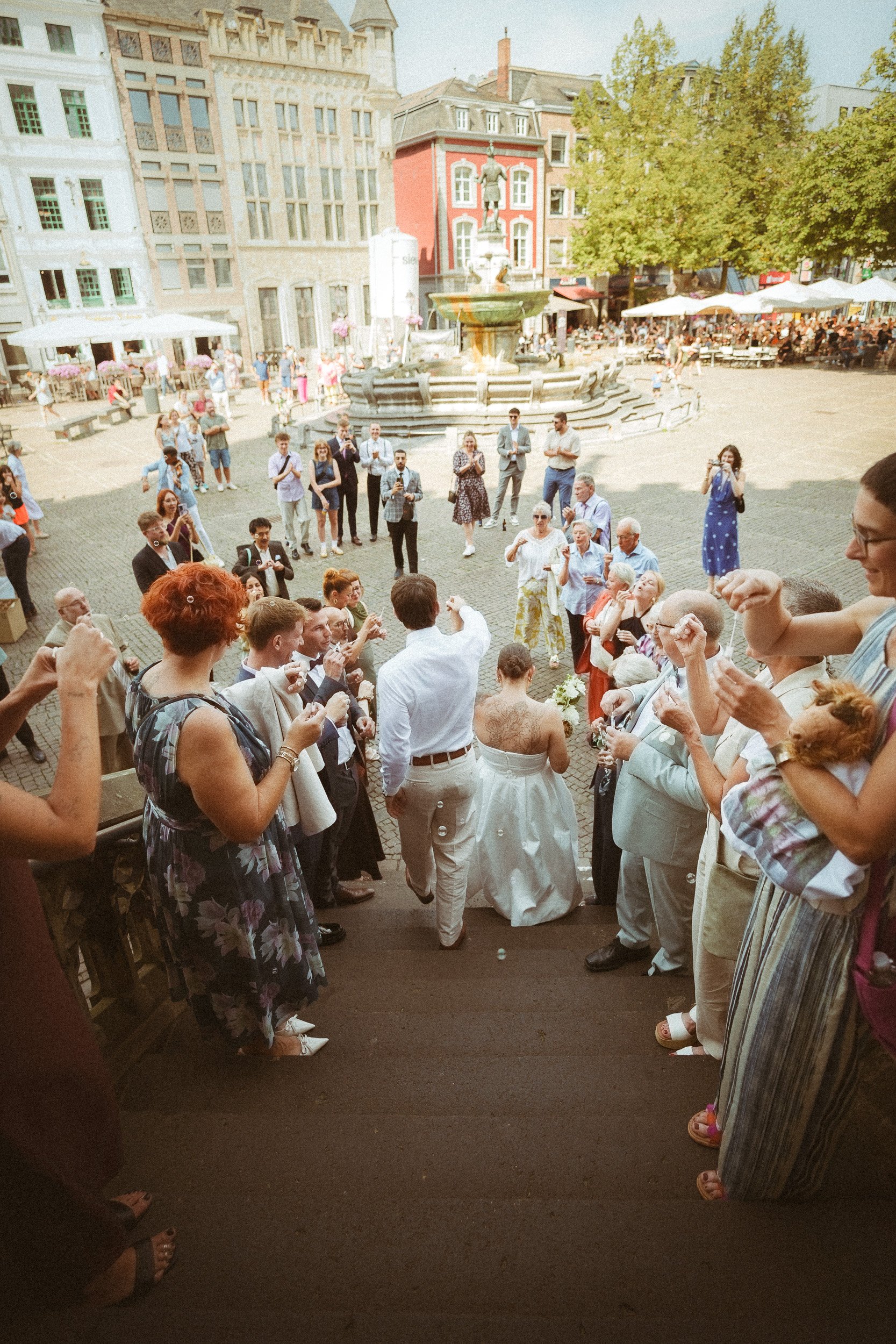 Hochzeit unter freiem Himmel vor einer barocken Stadtarchitektur mit Brunnen im Hintergrund, viele Gäste feiern und fotografieren das Brautpaar.