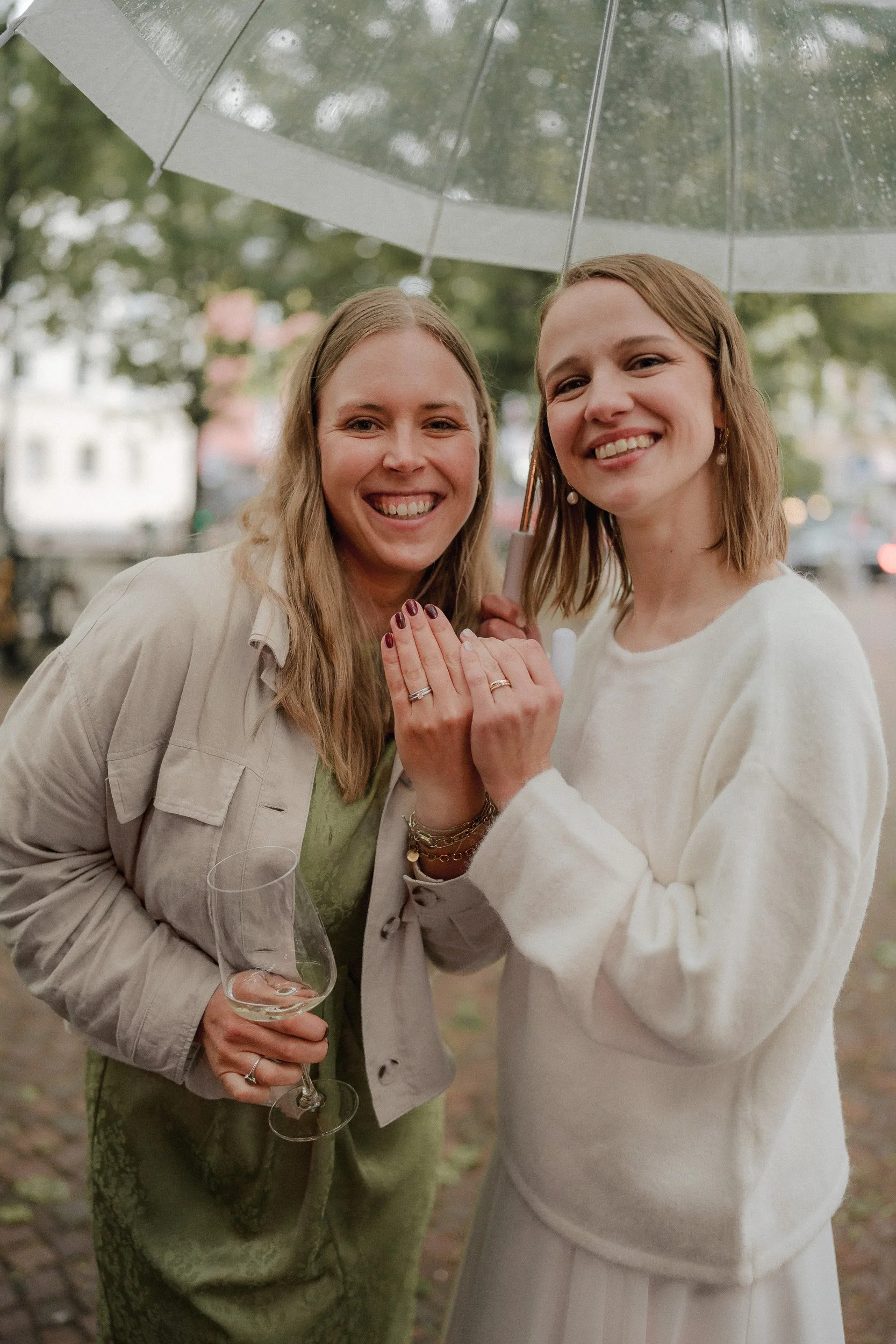 Zwei lachende Frauen unter einem transparenten Regenschirm im Freien, eine hält ein Weinglas, beide zeigen Eheringe.