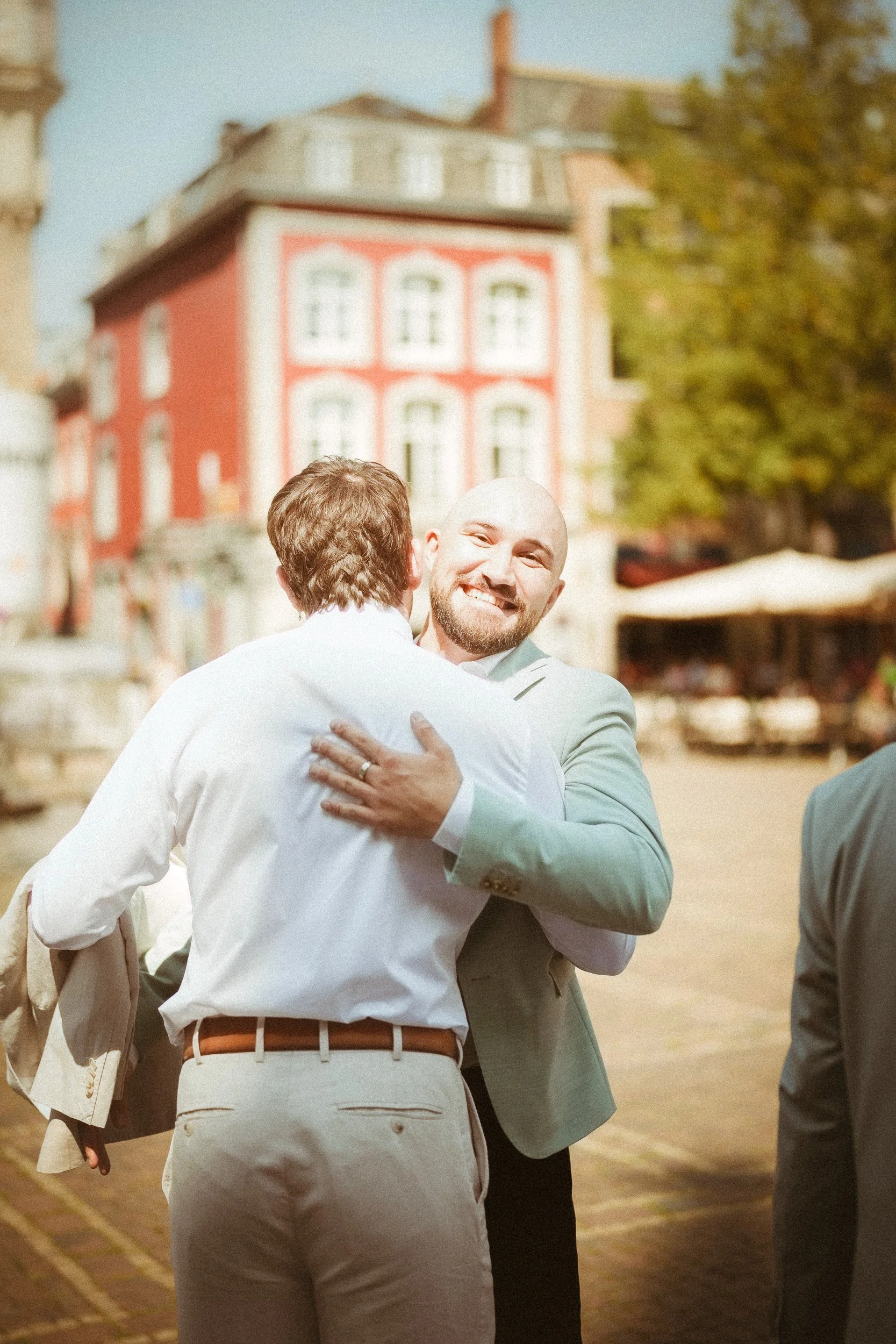 Zwei Männer umarmen sich und lächeln bei einem Treffen im Freien auf einer Straße, im Hintergrund bunte Gebäude und Bäume.