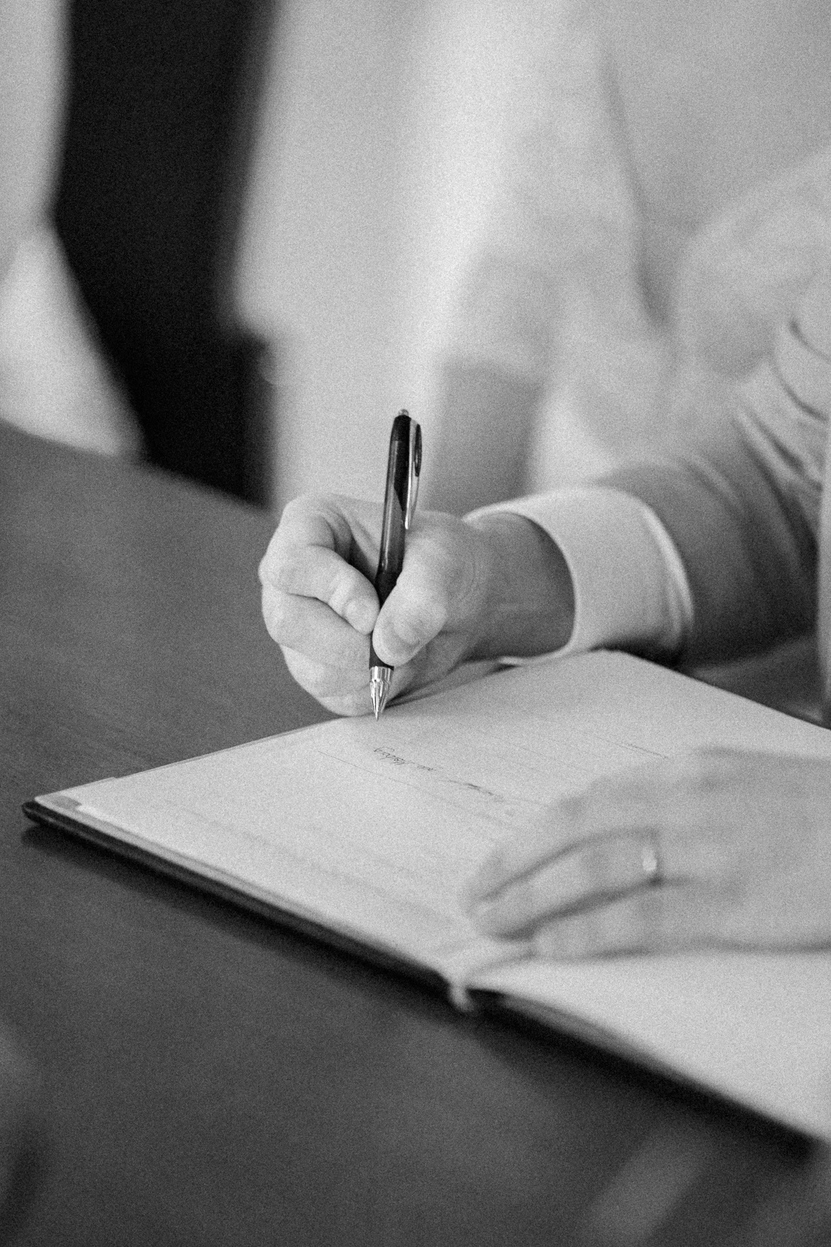 Schreibende Person trägt einen Anzug und schreibt in ein Notizbuch auf einem Holztisch.