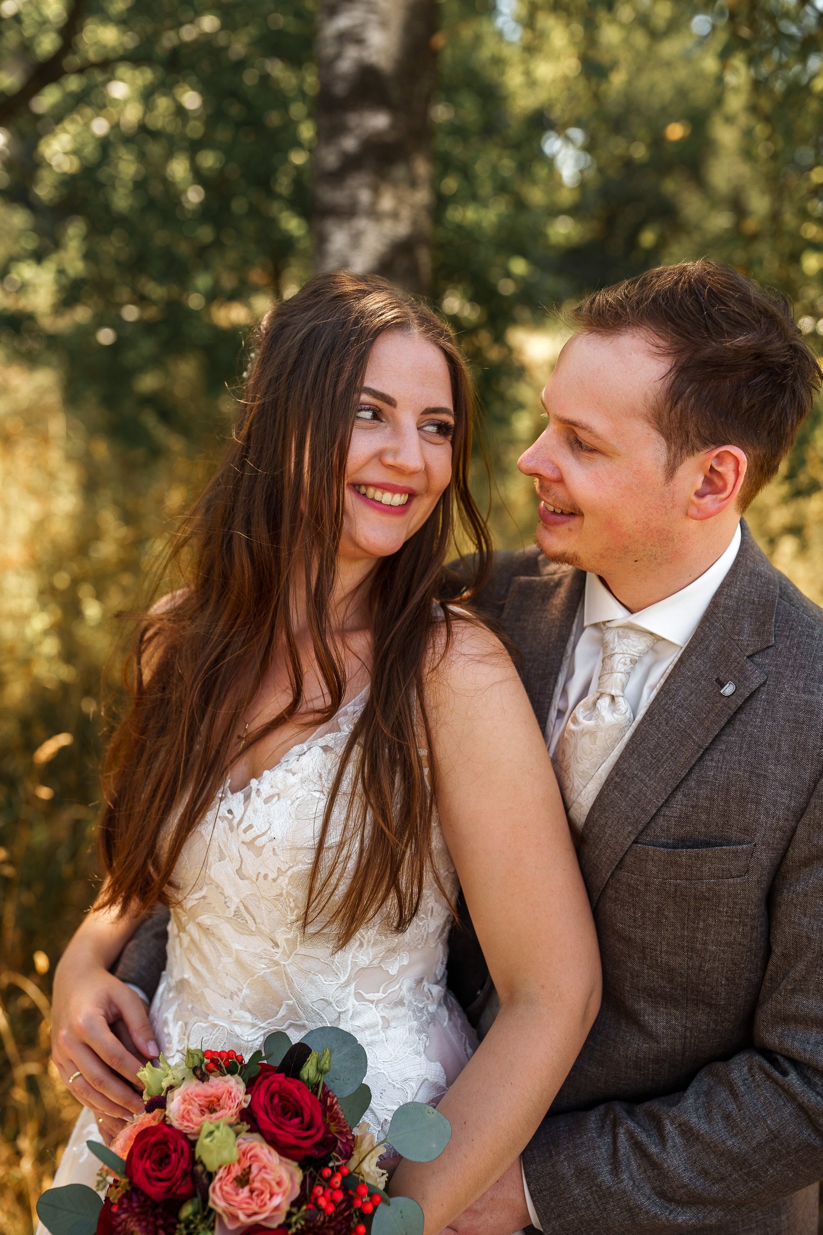 Ein glücklich wirkendes Brautpaar beim Fotoshooting im Freien, die Braut hält einen bunten Blumenstrauß, im Hintergrund Bäume.