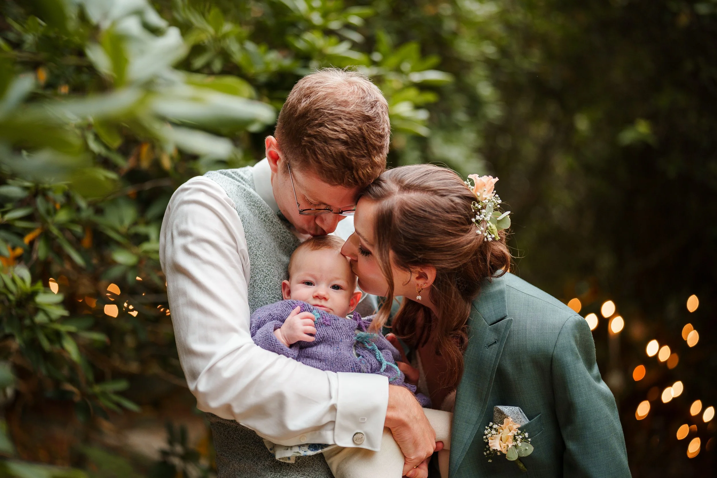 Ein Mann und eine Frau, die ein Baby halten, küssen sich gegenseitig bei einer Hochzeit im Freien. Die Frau trägt einen grünen Anzug mit Blumen im Haar, der Mann trägt eine Weste, und im Hintergrund sind Lichter und grüne Bäume sichtbar.