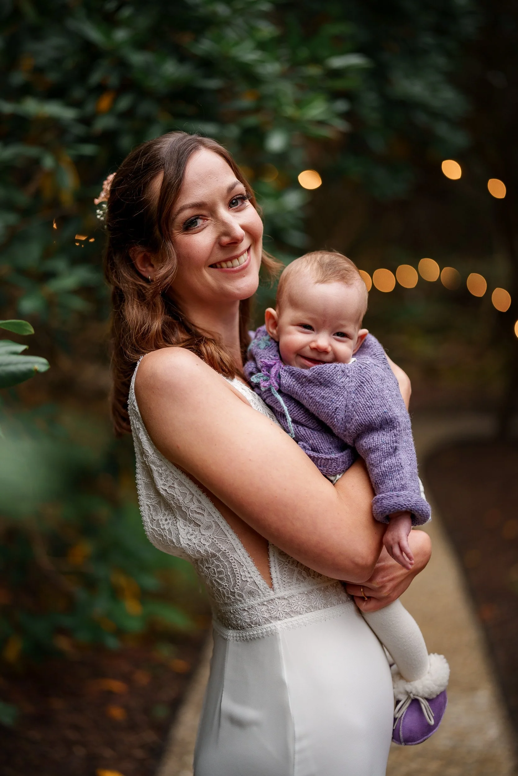 Eine lachende Frau in einem weißen Kleid hält ein lächelndes Baby in einem lila Pullover und weißen Strumpfhosen vor einem grünen Hintergrund mit Lichterketten.