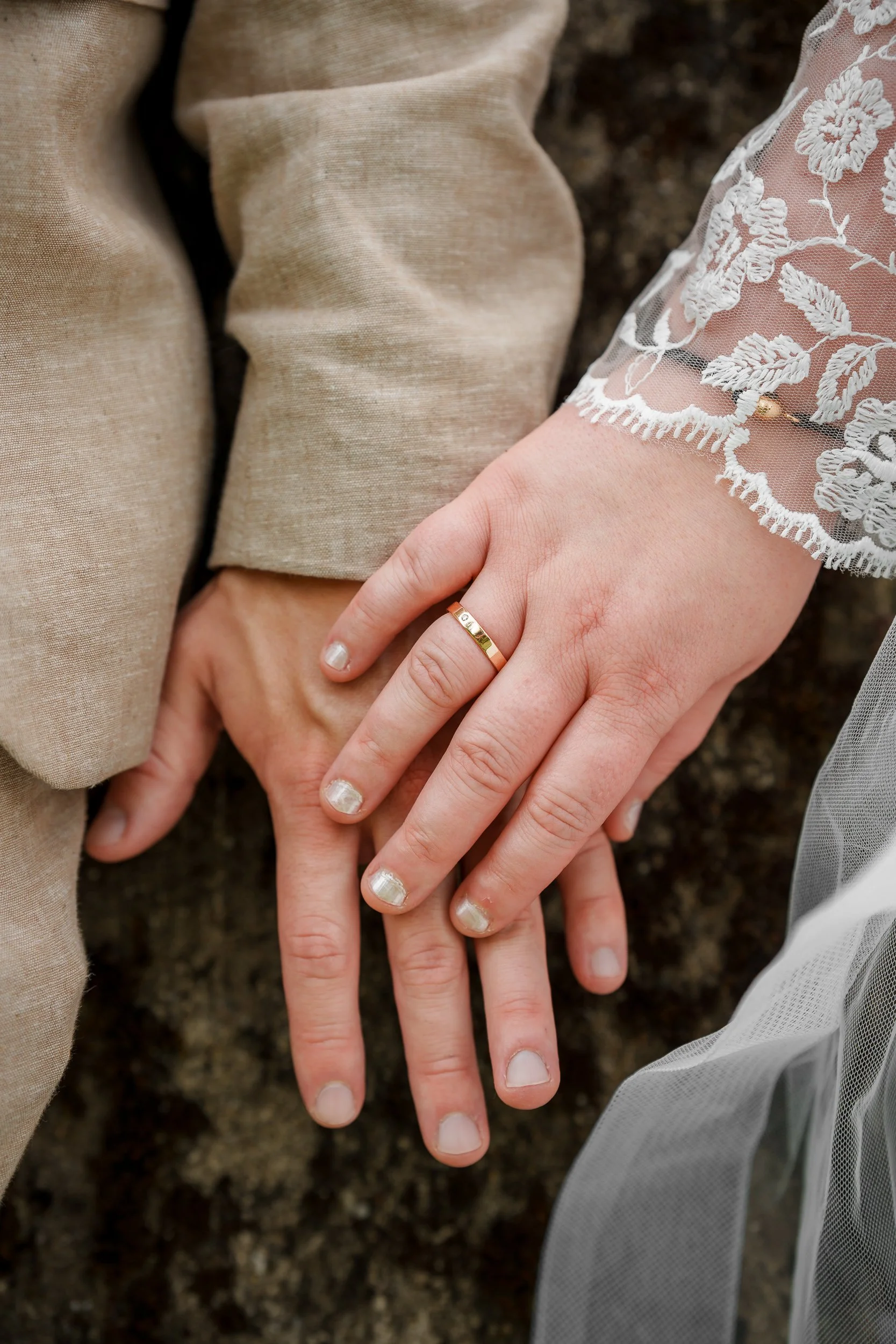 Nahaufnahme von zwei Händen, die auf dem Boden liegen, die eine trägt einen Ehering, die andere eine helle, lange Spitze mit Stickerei. 