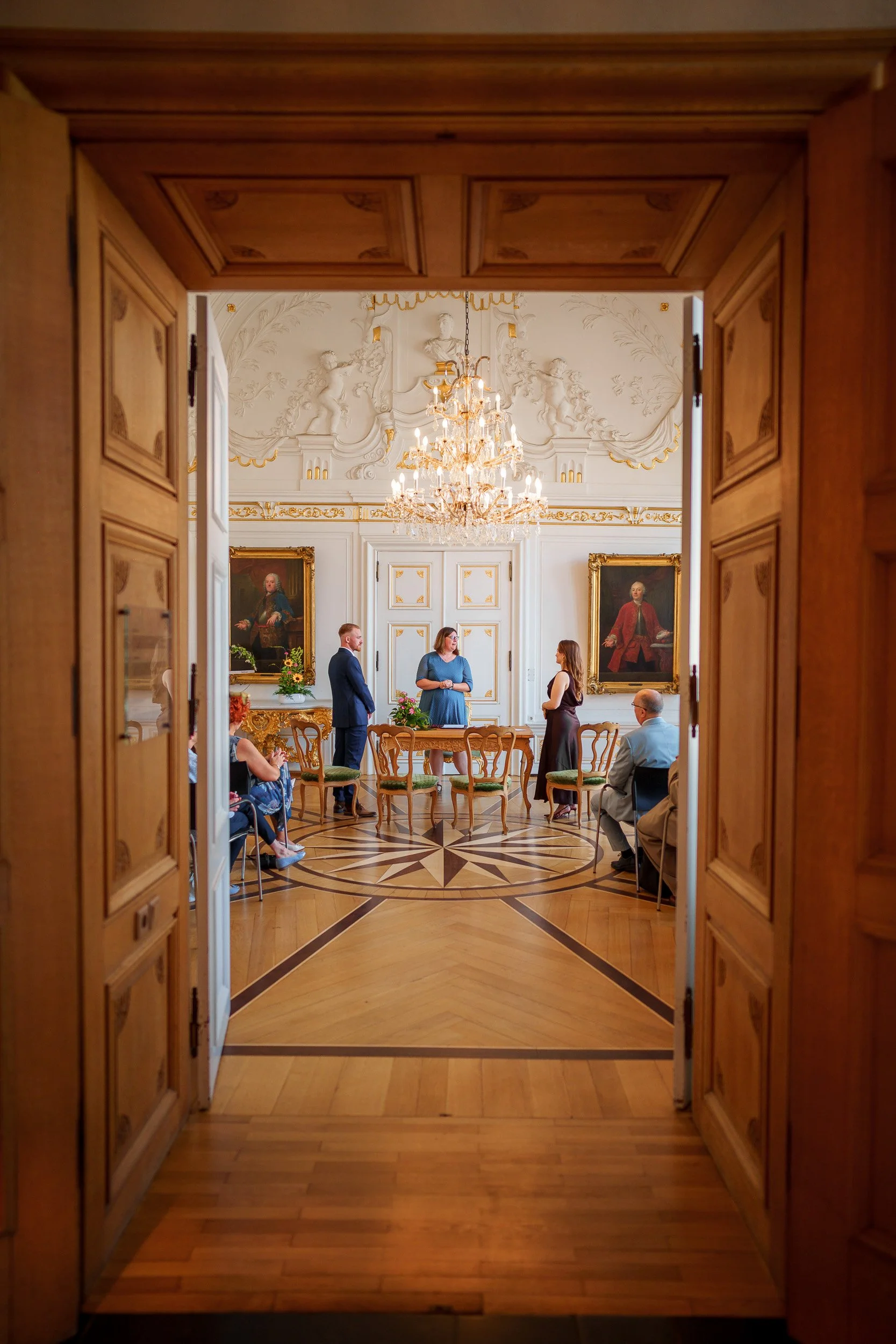 Blick in einen eleganten, prunkvollen Saal durch eine offene Tür. Im Raum sind mehrere Personen, die sich unterhalten, umgeben von barockem Dekor, Marmorpfeilern, Gemälden und einem Kristallleuchter.