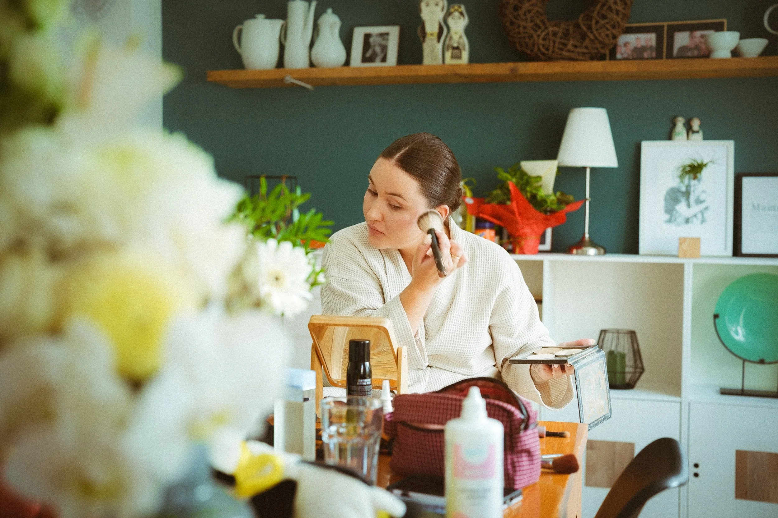 Junge Frau trägt weißen Bademantel und schminkt sich mit Puderpinsel vor einem Tisch mit Kosmetikartikeln und Spiegel.